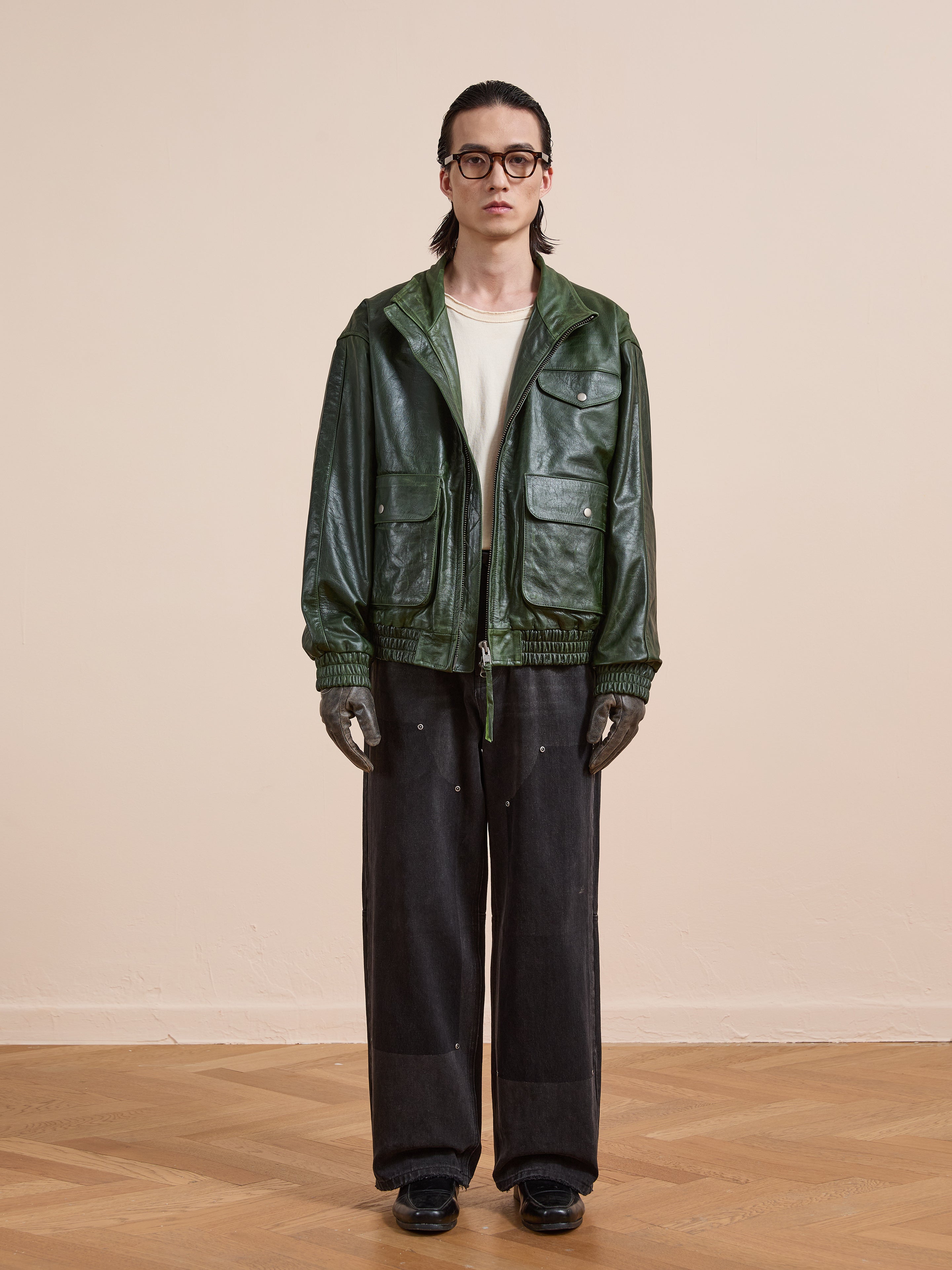 A person stands indoors on a wooden floor against a beige wall, wearing glasses, gloves, black shoes, dark wide-leg pants, a white shirt, and the FOUND Forest Leather Bomber Jacket.