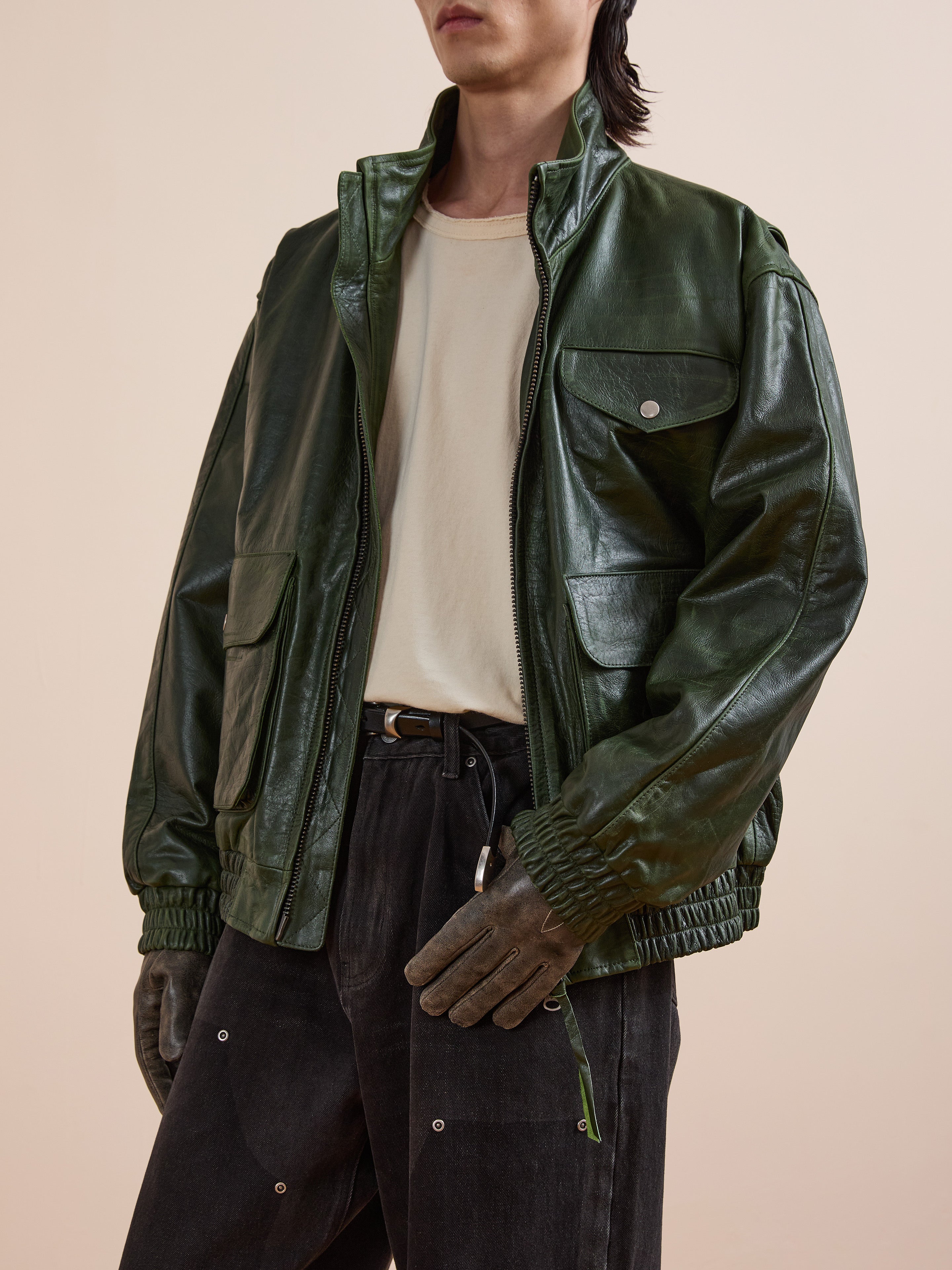 Wearing FOUND’s Forest Leather Bomber Jacket, an off-white tee, dark jeans with metal accents, and green gloves, a person stands against a plain beige background.