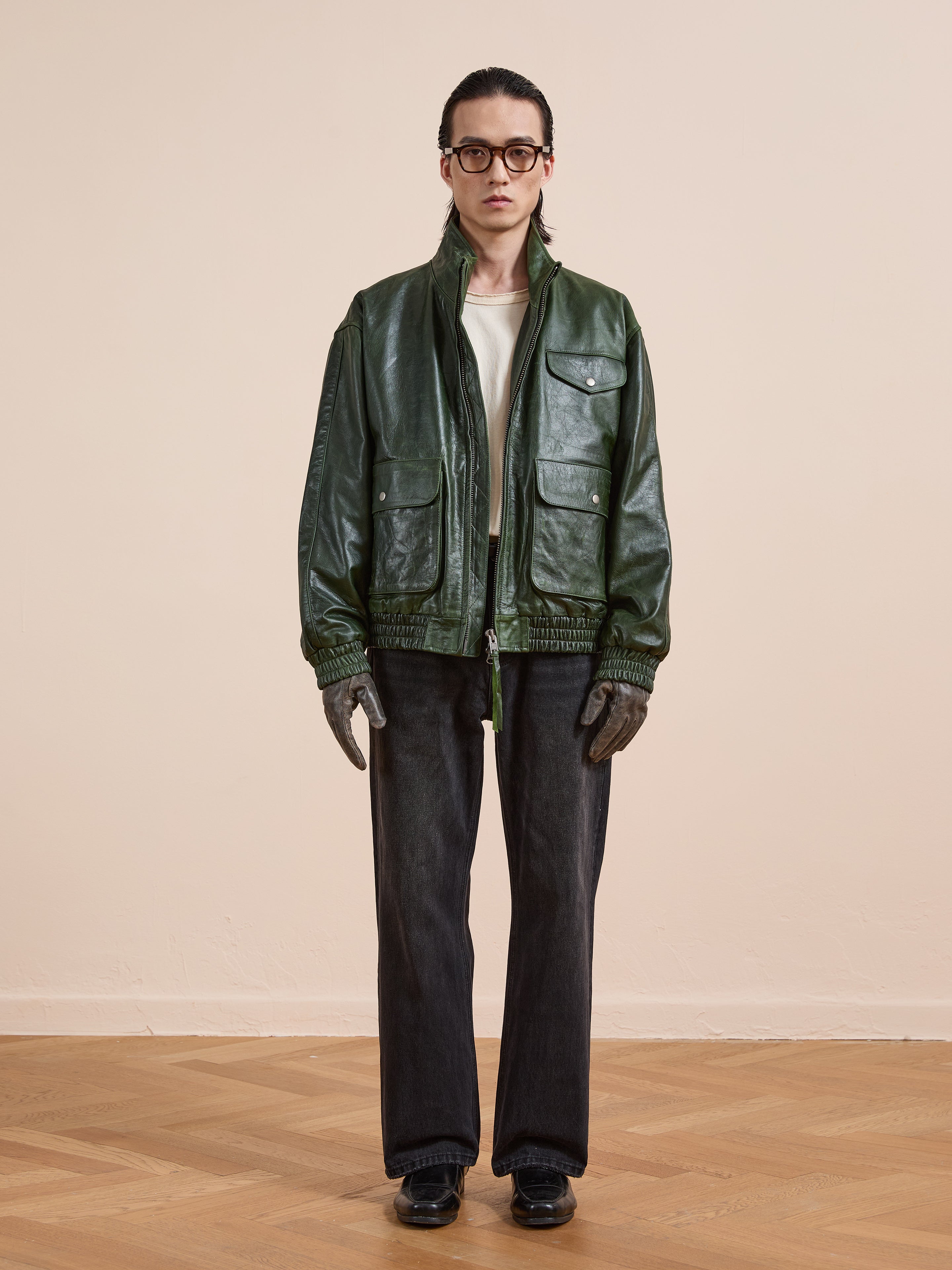 A person stands indoors against a plain beige wall wearing FOUND’s Forest Leather Bomber Jacket, glasses, a white shirt, dark jeans, gloves, and black shoes.