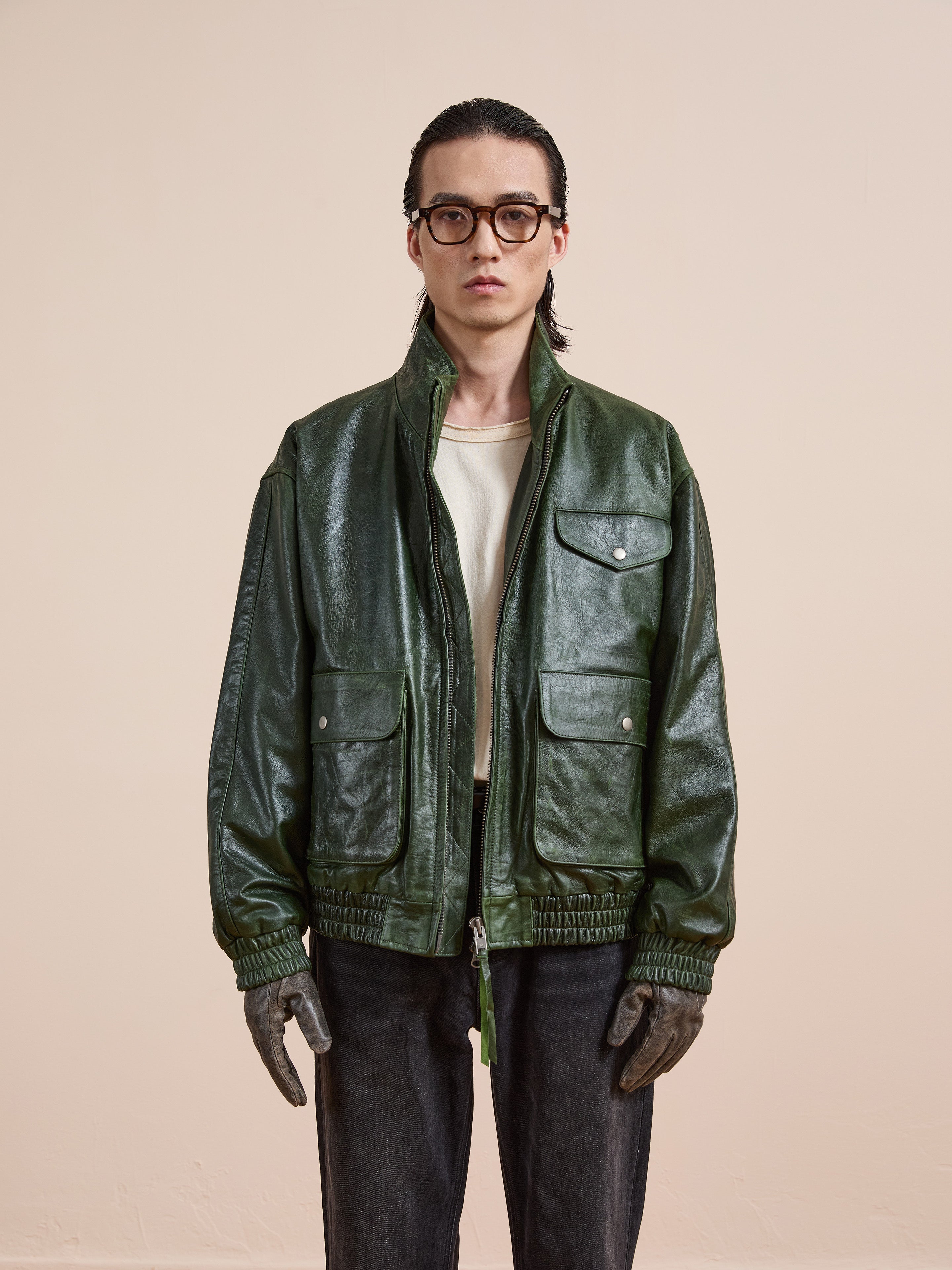 A person in a FOUND Forest Leather Bomber Jacket, white shirt, dark pants, gray gloves, and eyeglasses stands against a plain beige background.