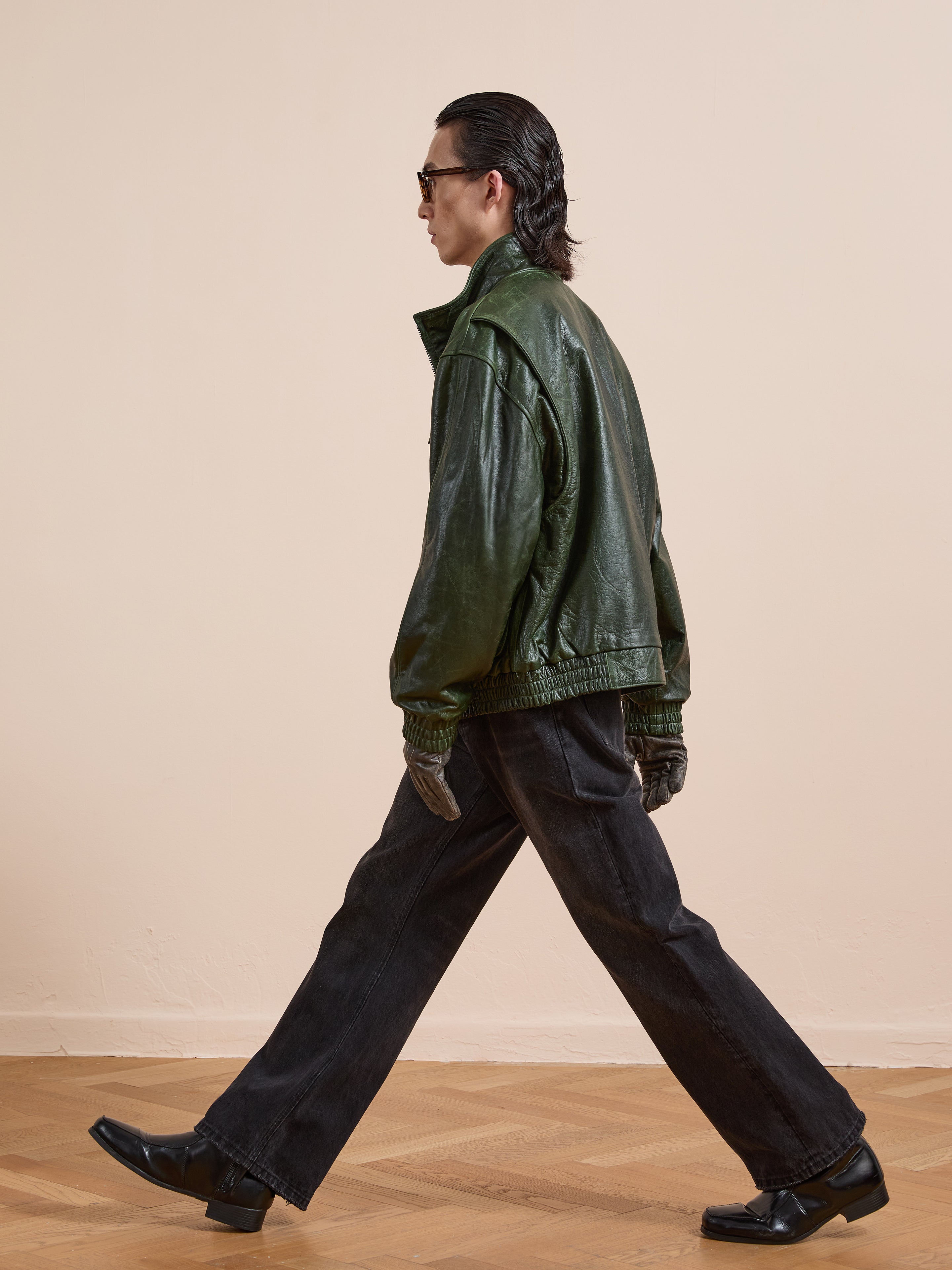 A person wearing FOUND’s Forest Leather Bomber Jacket, sunglasses, dark jeans, and black shoes walks indoors on a parquet floor against a beige wall.