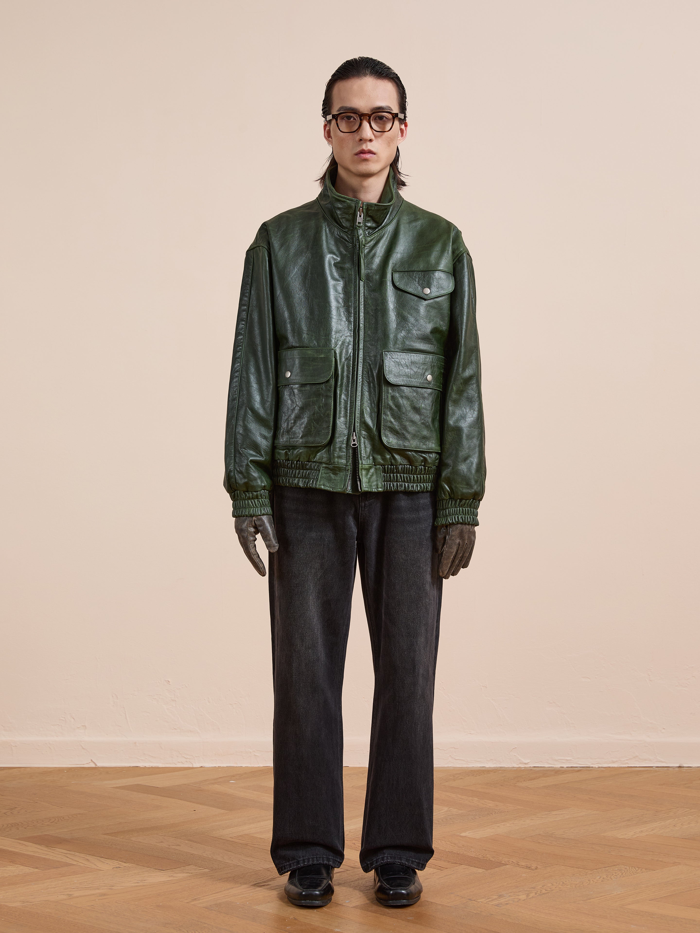 A person stands indoors against a beige wall wearing the FOUND Forest Leather Bomber Jacket, dark gray pants, black shoes, gray gloves, and round glasses.