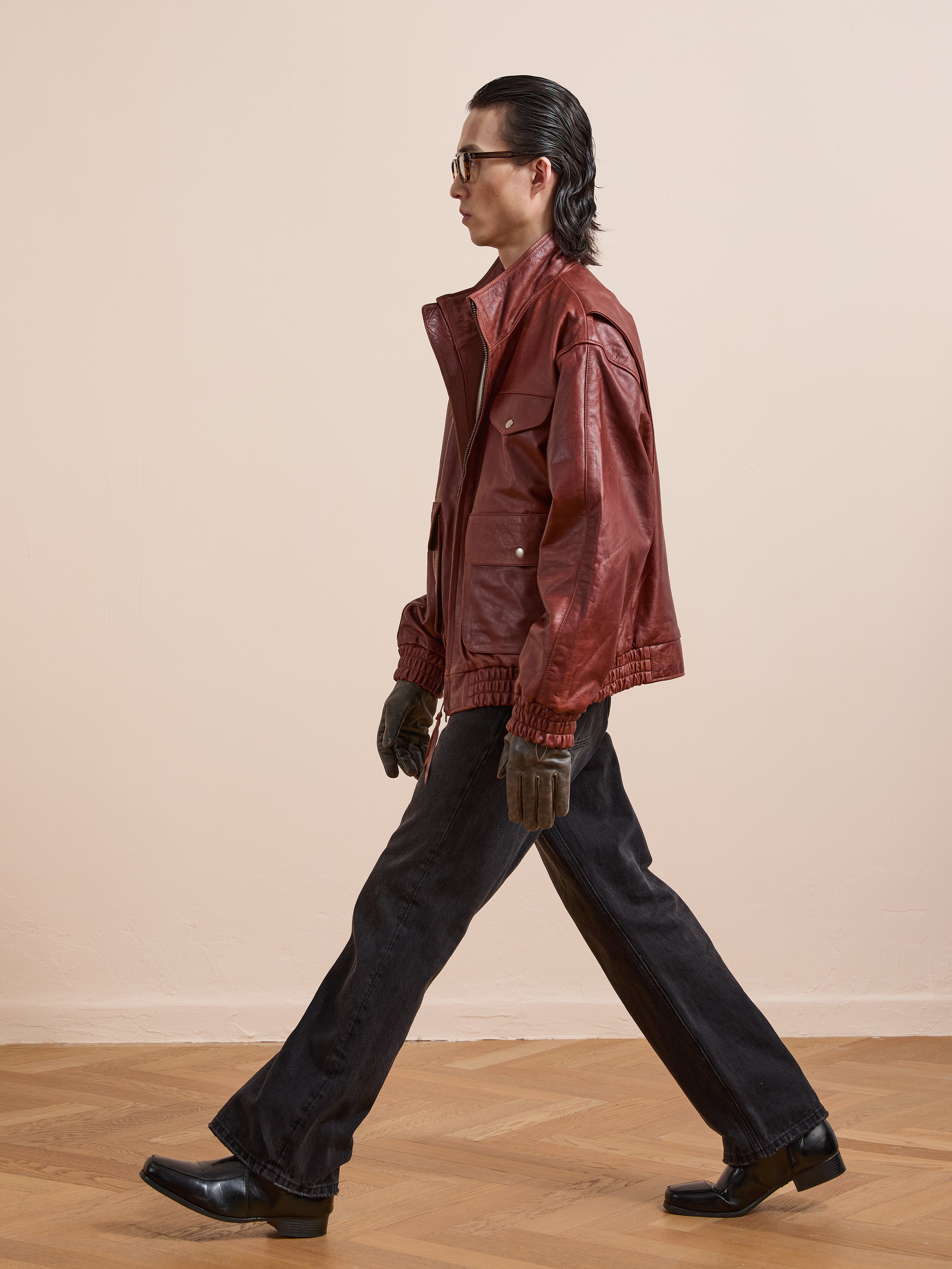 Wearing the Found Redwood Leather Bomber Jacket, black flared pants, gloves, and boots, a person walks across a wooden floor against a plain beige backdrop.