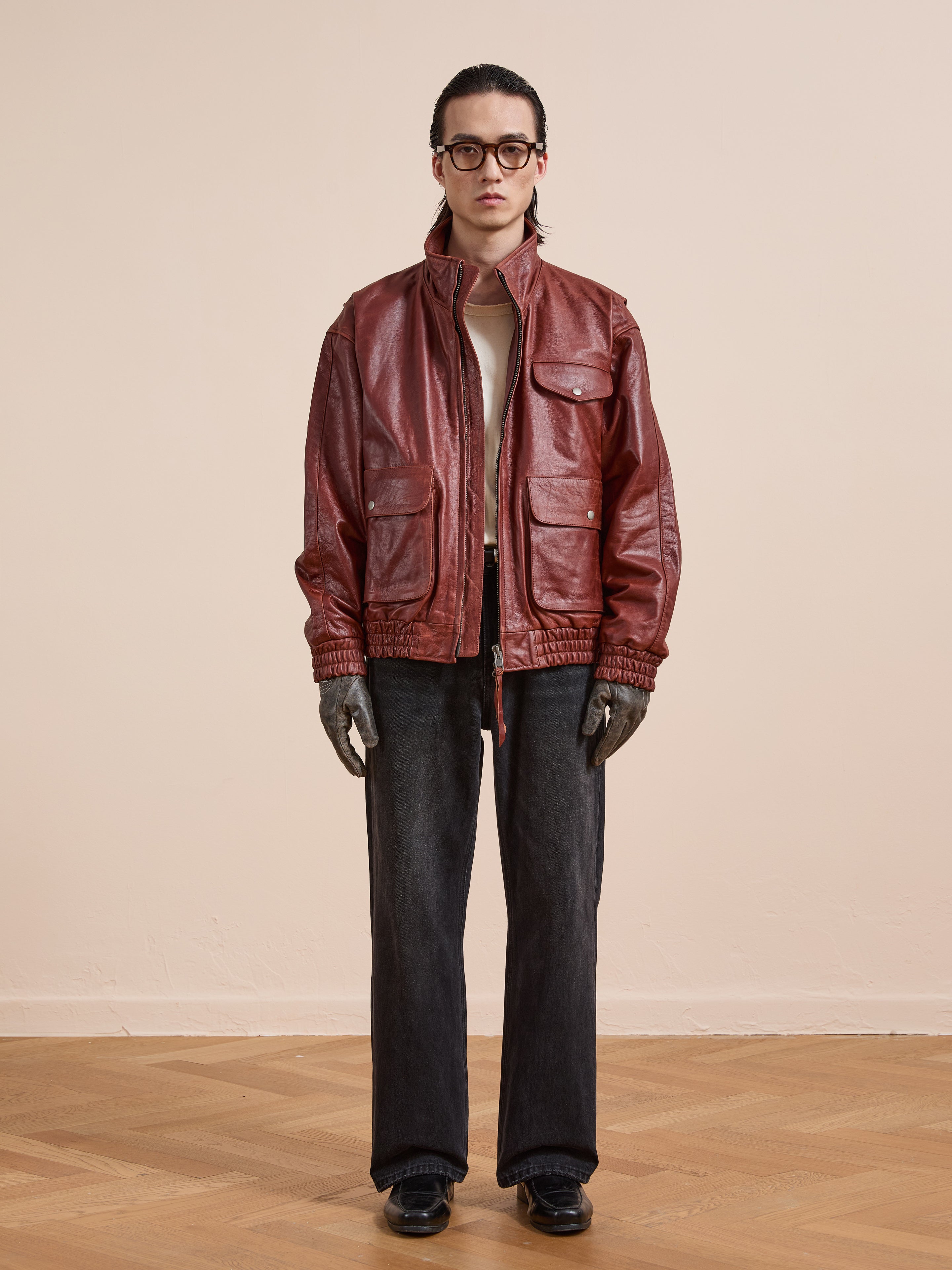 A person stands indoors against a beige wall and wooden floor, wearing the Found Redwood Leather Bomber Jacket, an off-white shirt, black wide-leg jeans, black shoes, gray gloves, and glasses.