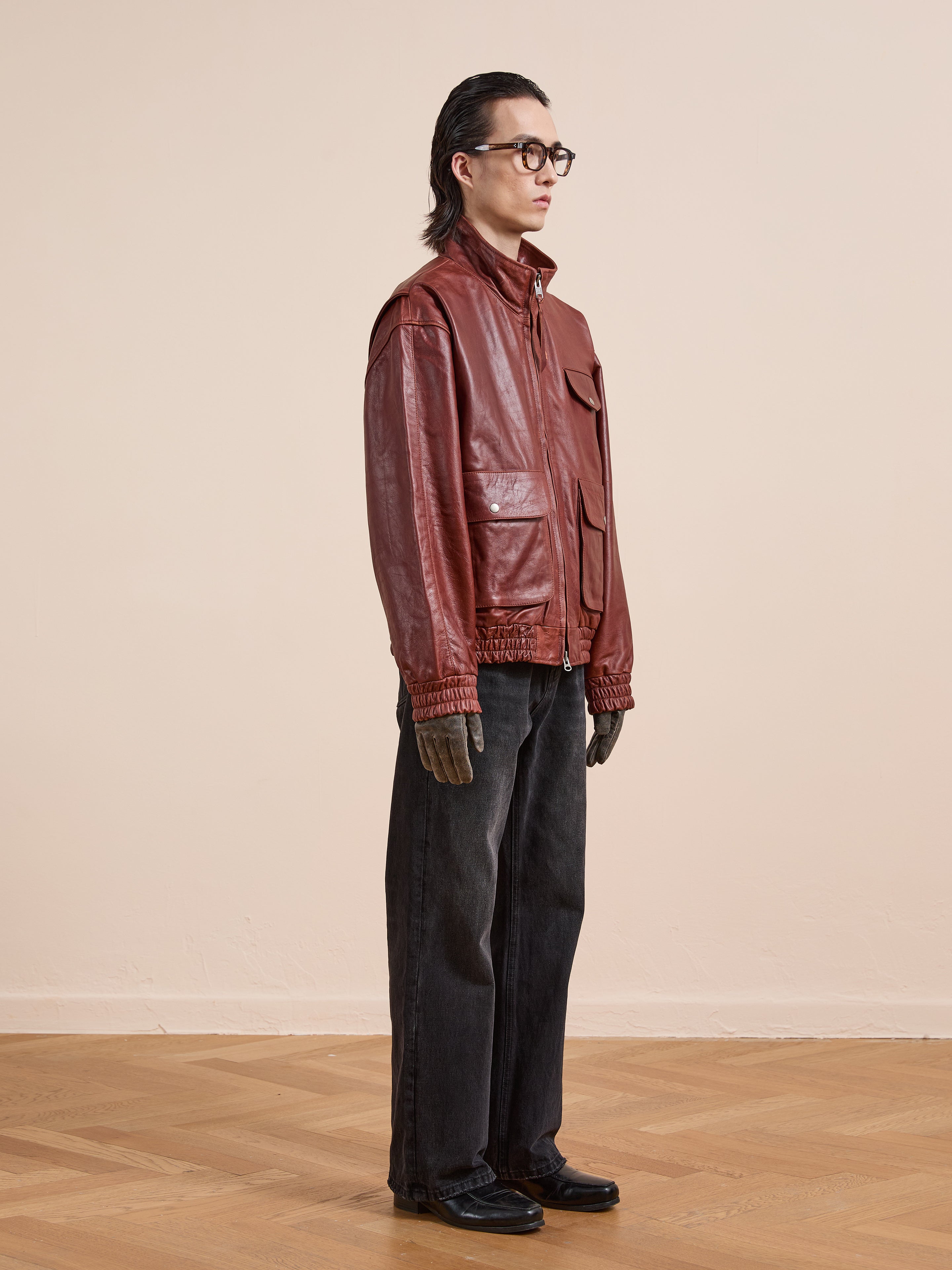 Indoors, a person wears the Found Redwood Leather Bomber Jacket in red with dark wide-leg jeans, black shoes, brown gloves, and glasses against a plain beige wall and wooden floor.