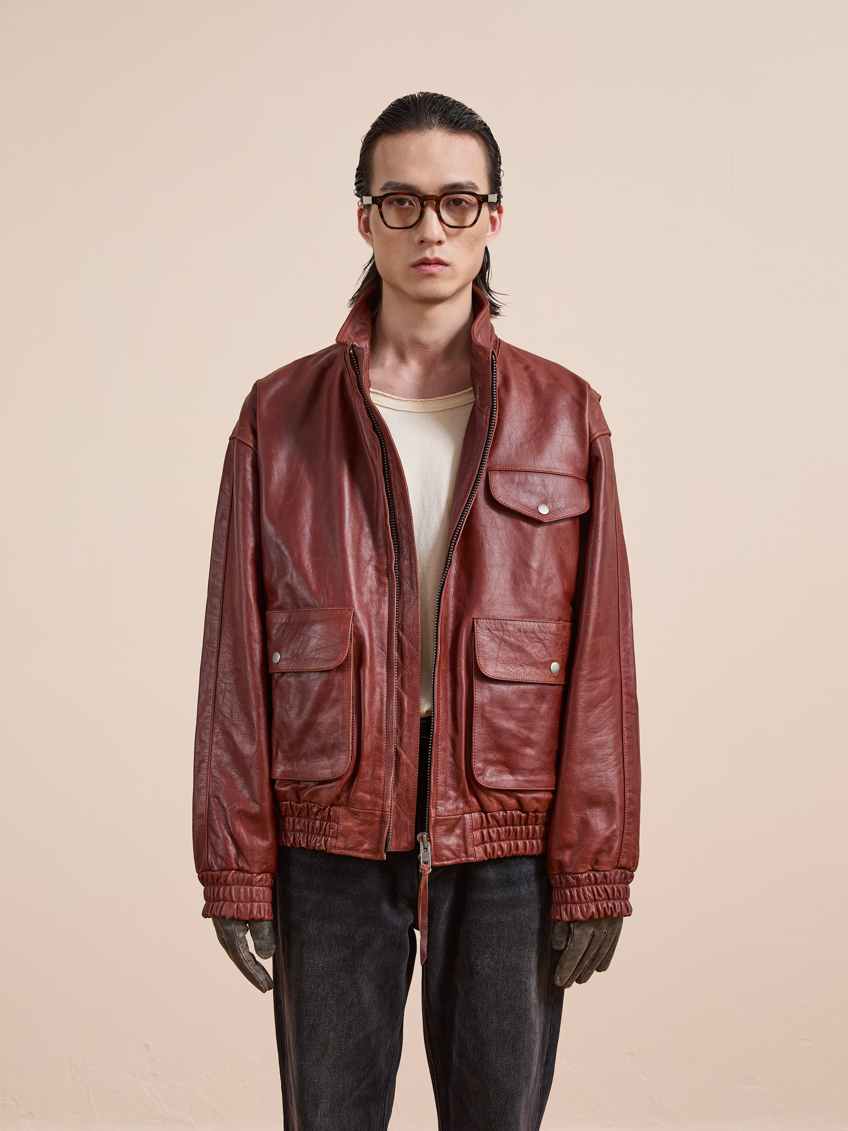 A person with long hair and glasses wears the Found Redwood Leather Bomber Jacket over a beige shirt, paired with dark jeans and gray gloves, standing against a plain light background.