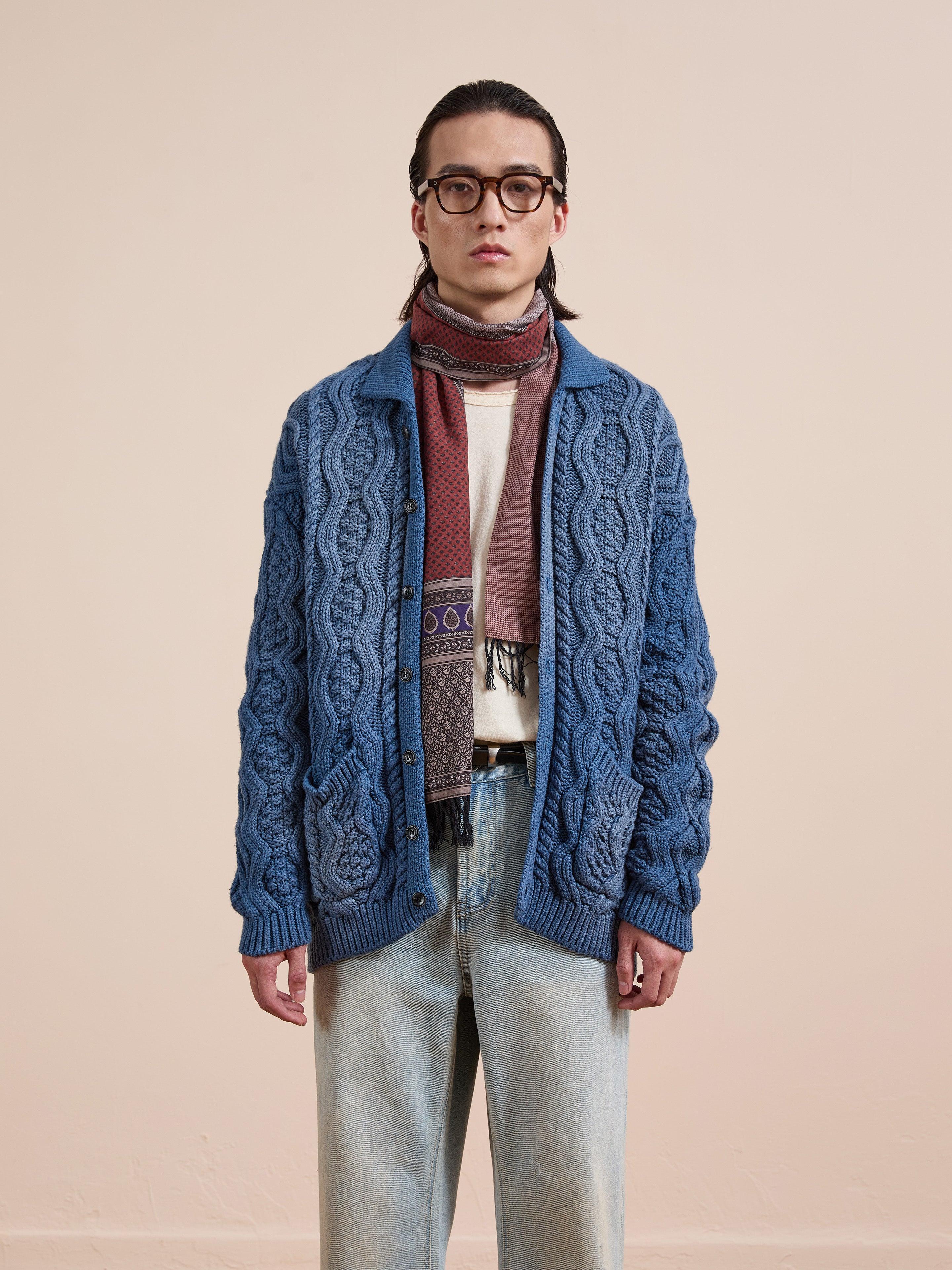 A person in Found's Cable Knit Buttoned Cardigan, an off-white shirt, light jeans, a patterned scarf, and glasses stands against a plain beige background.