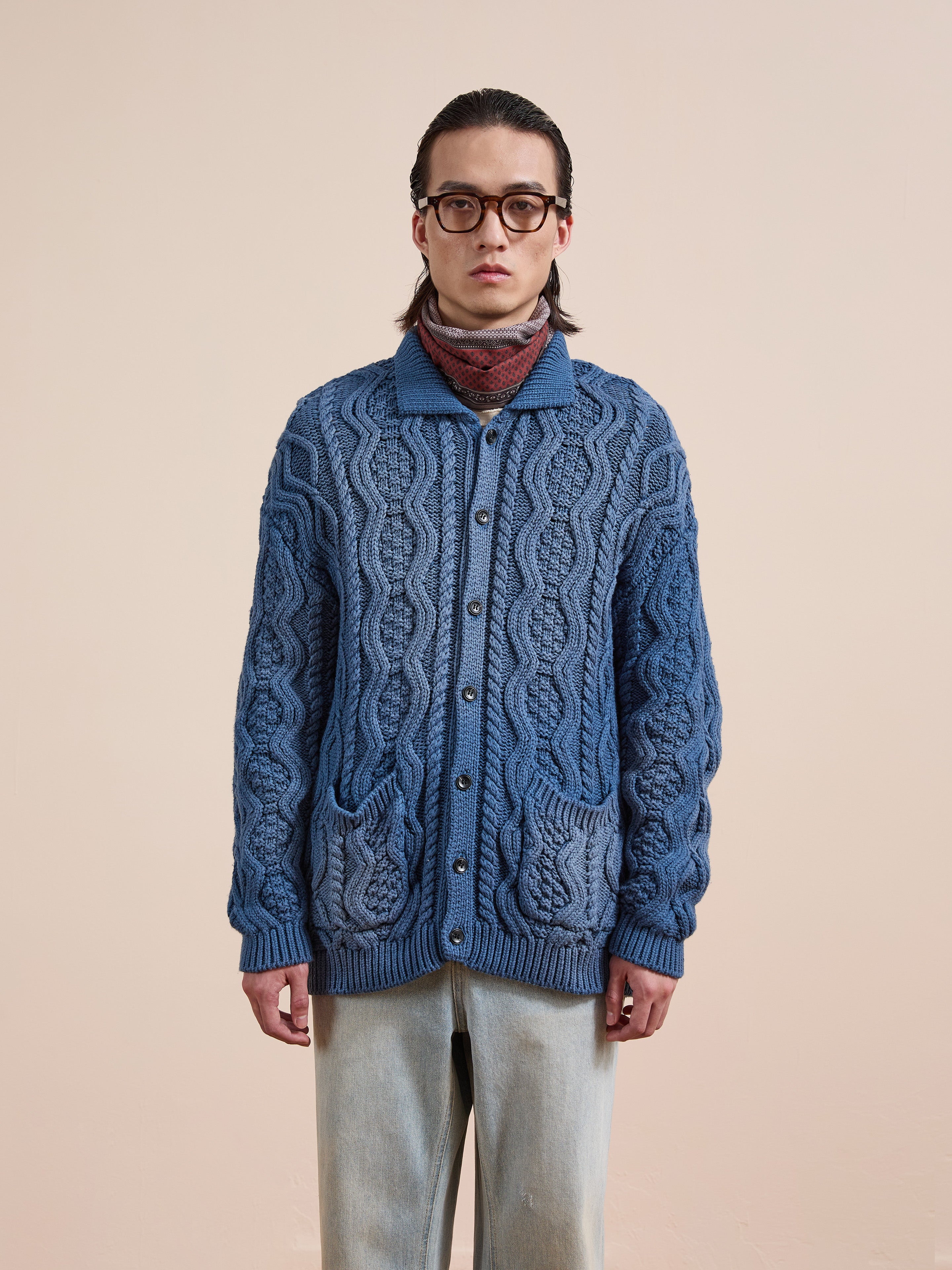 A person wearing glasses, the Found Cable Knit Buttoned Cardigan in navy, a red patterned scarf, and light-wash jeans stands against a plain beige background.