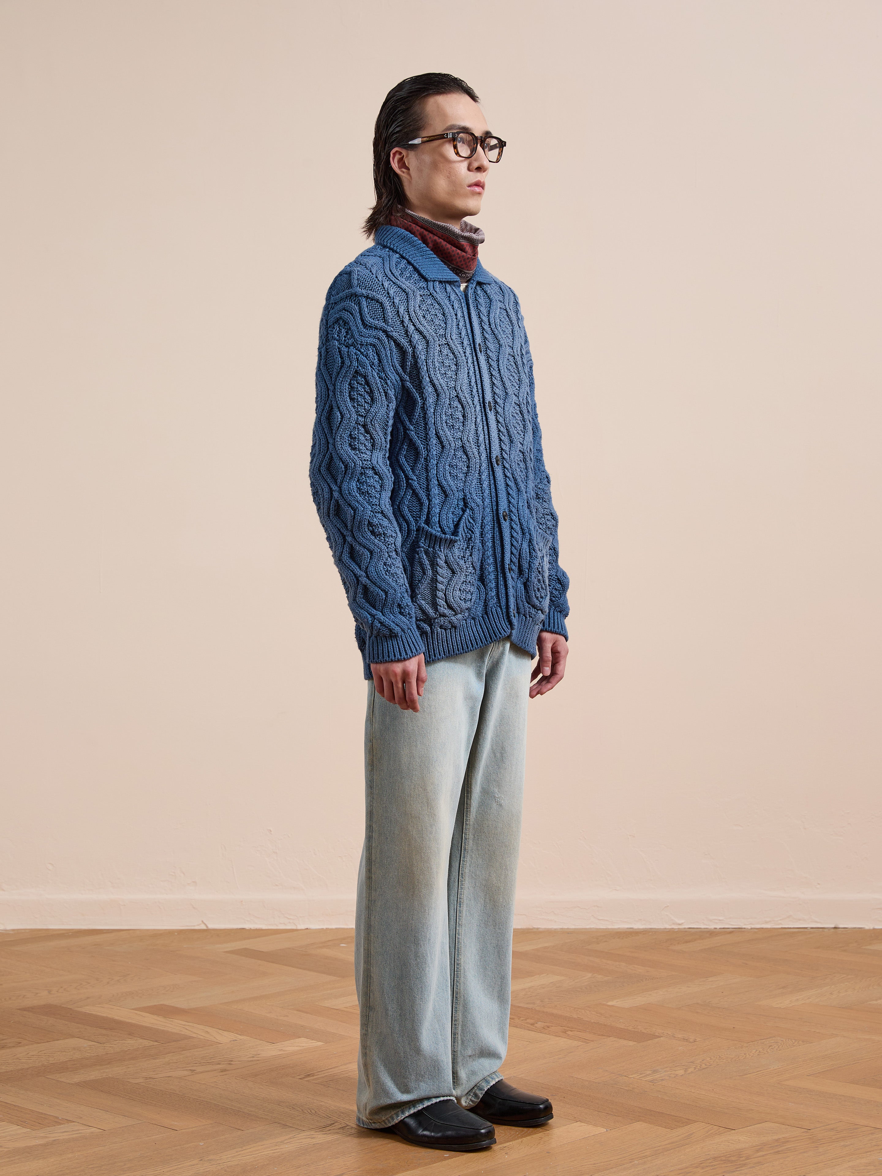 A person wears the Found Cable Knit Buttoned Cardigan in navy with light-wash wide-leg jeans, black shoes, glasses, and a brown scarf, standing indoors against a plain beige wall and hardwood floor.