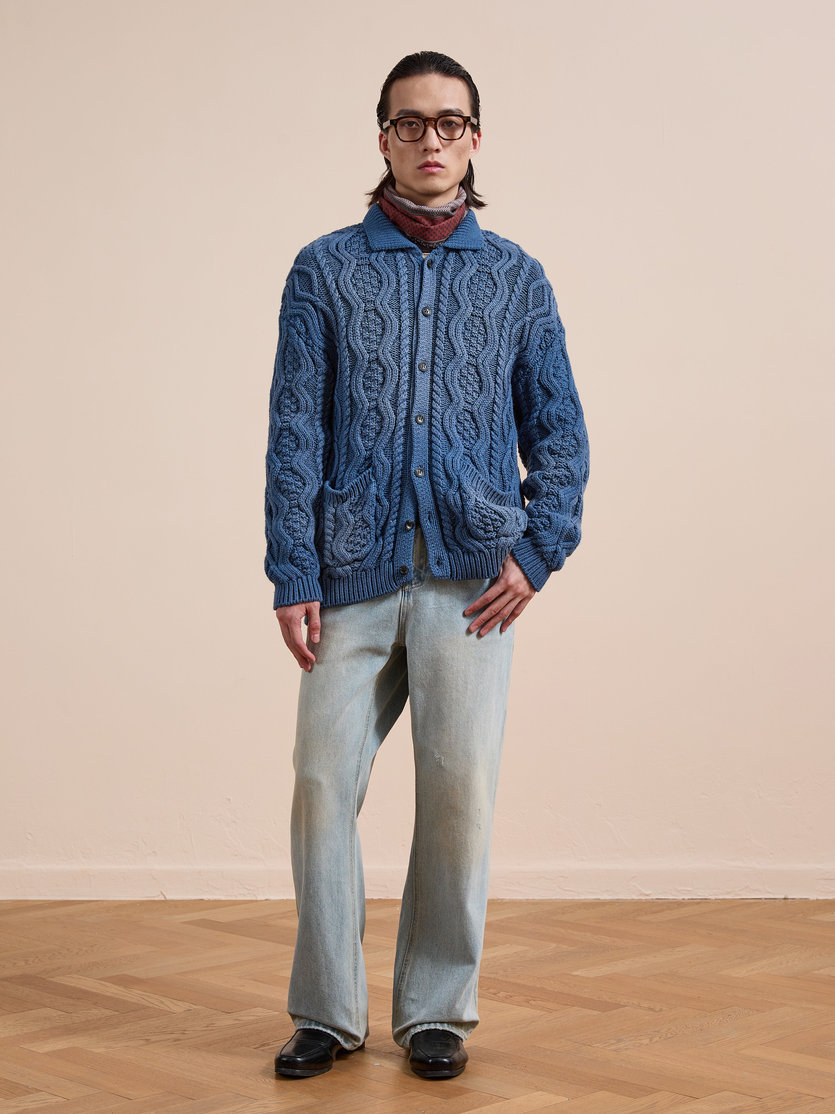 A person wearing Found’s Cable Knit Buttoned Cardigan in blue, with light blue jeans, black shoes, glasses, and a red scarf stands on a wooden floor against a plain beige wall. This classic piece offers timeless unisex style.