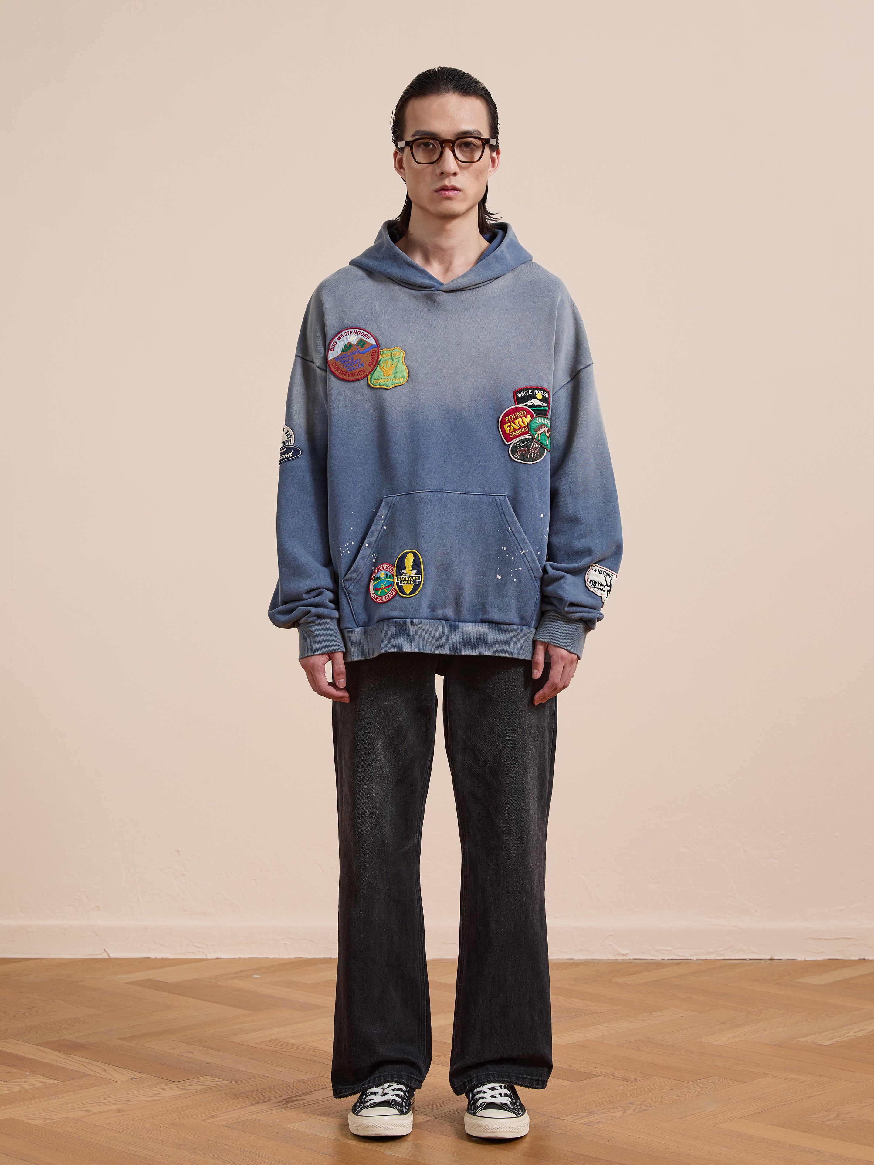 A person wearing black glasses, a FOUND Distressed Patch Hoodie, dark jeans, and black-and-white sneakers stands on a wooden floor against a plain beige wall.