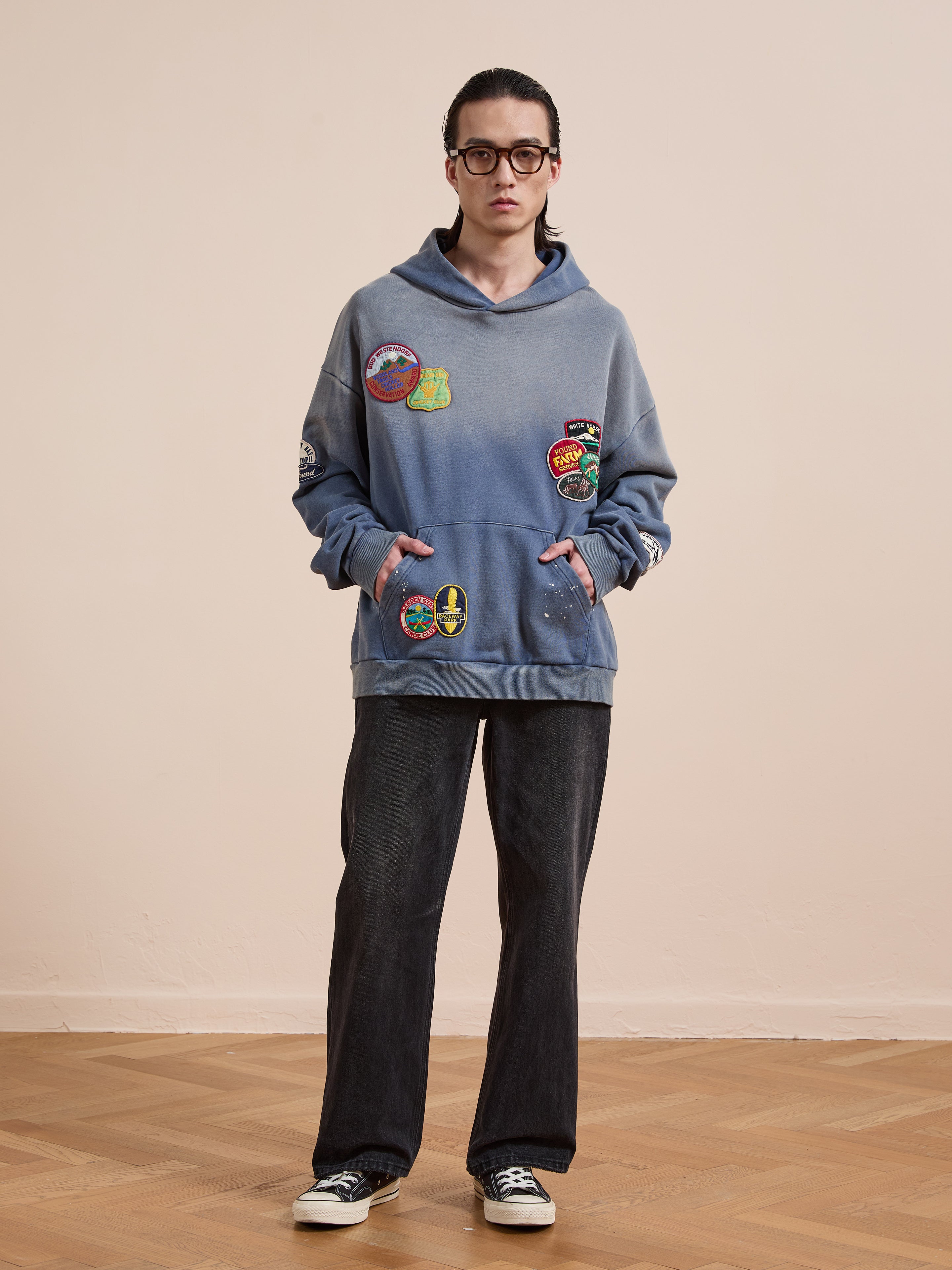 Person standing indoors on a parquet floor, wearing glasses, the FOUND Distressed Patch Hoodie, black wide-leg jeans, and black-and-white sneakers.