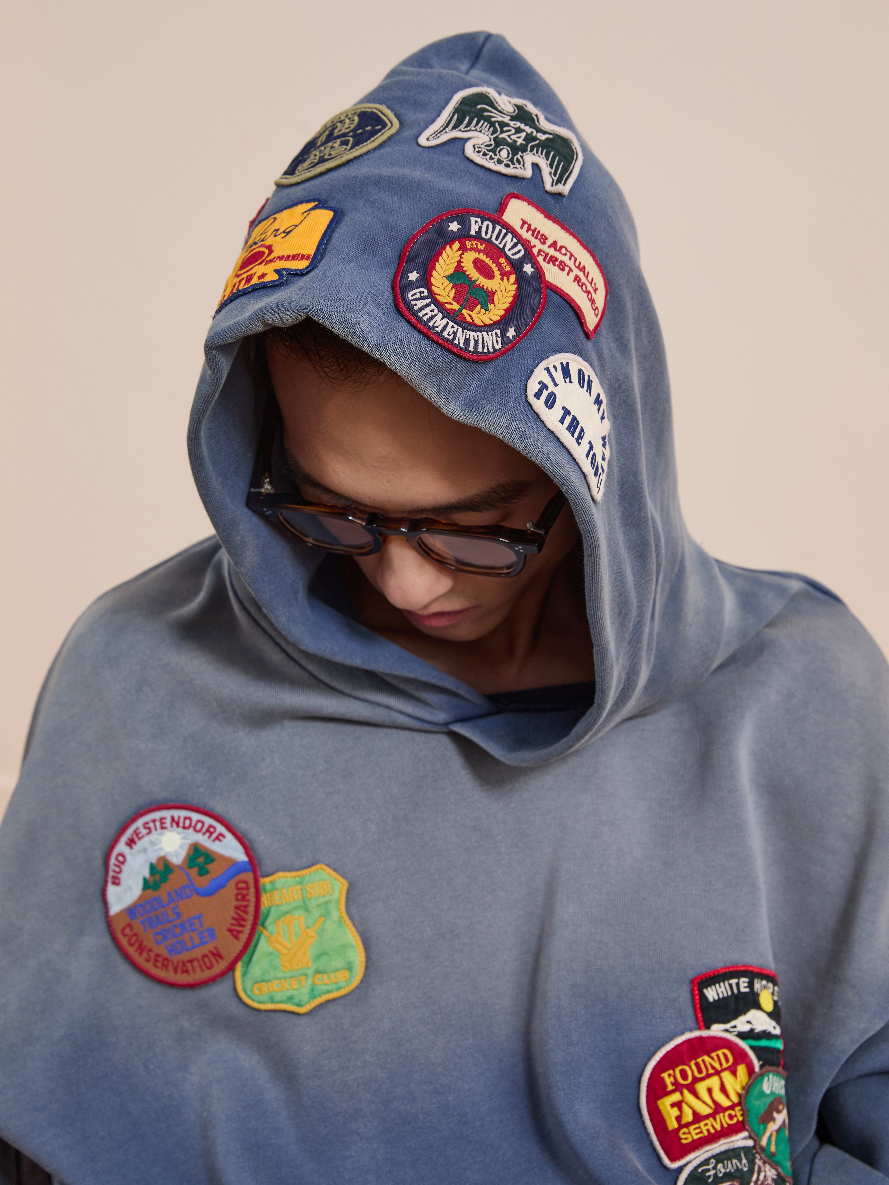 A person wearing sunglasses and the FOUND Distressed Patch Hoodie in blue French Terry cotton, embellished with colorful embroidered patches, looks downward.