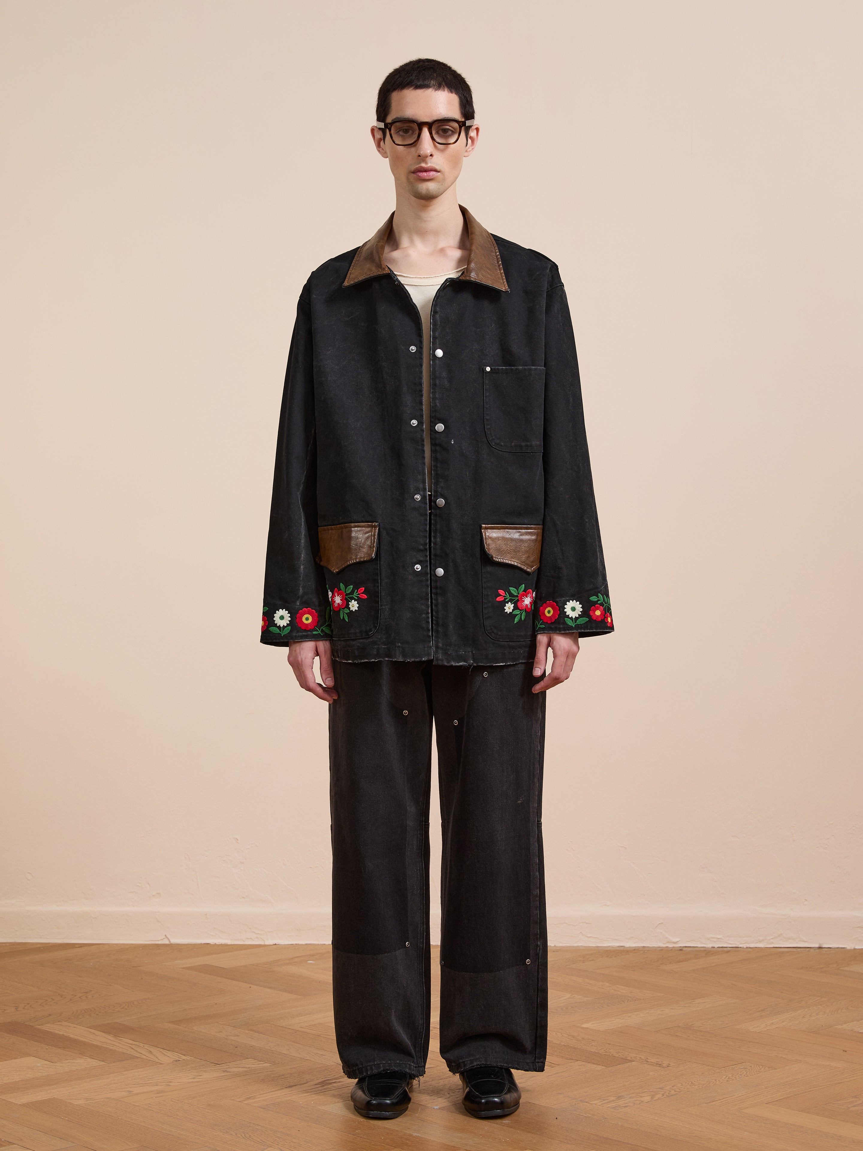 Indoors, a person wears the Found Canvas Embroidered Work Jacket—unisex, relaxed fit, black with brown accents and floral embroidery on the cuffs—styled with wide-leg pants and black shoes.