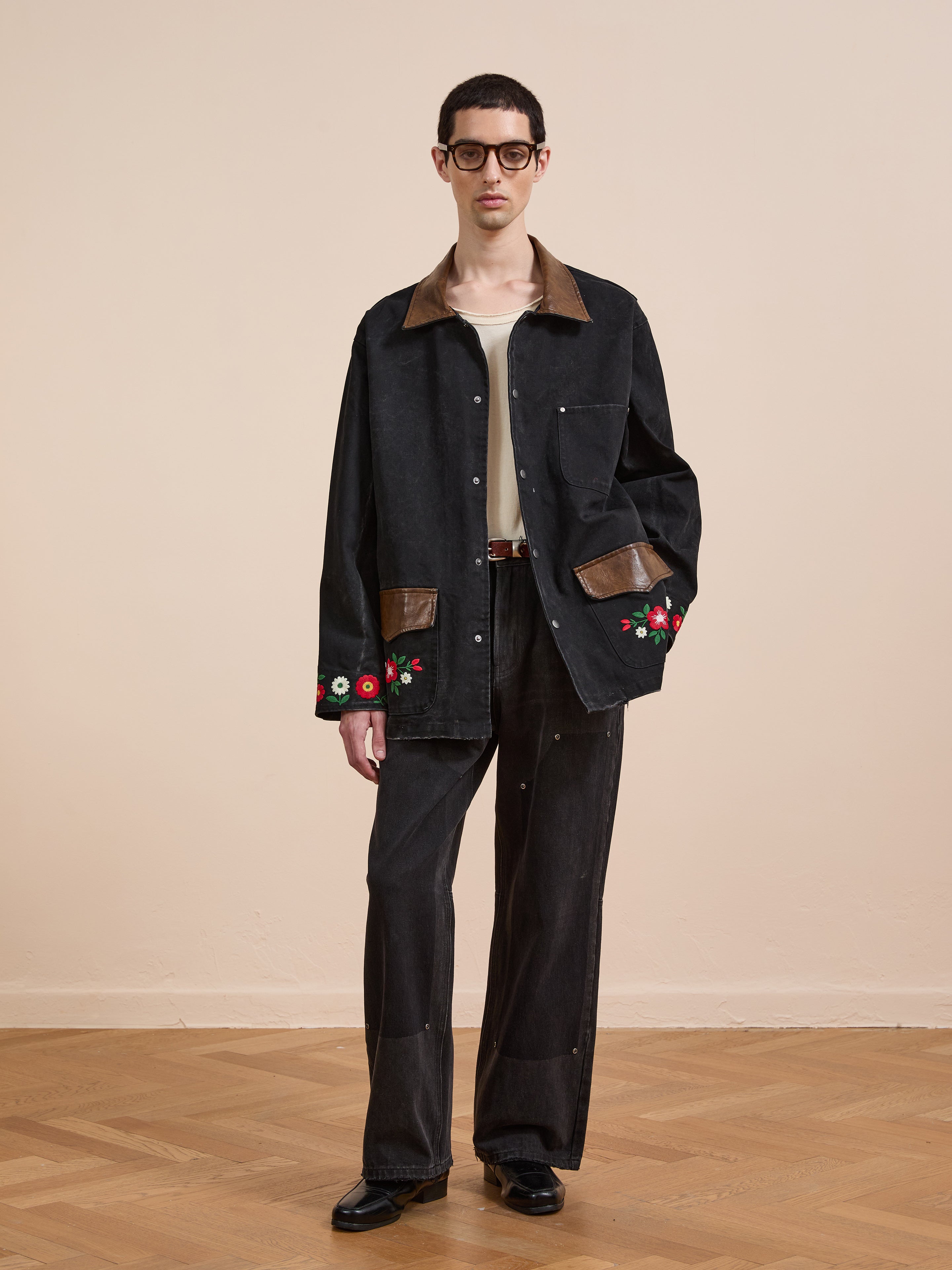 A person wearing glasses, Found’s Canvas Embroidered Work Jacket with brown accents and floral embroidery, a beige shirt, dark wide-leg pants, and black shoes stands on wooden flooring against a plain background.