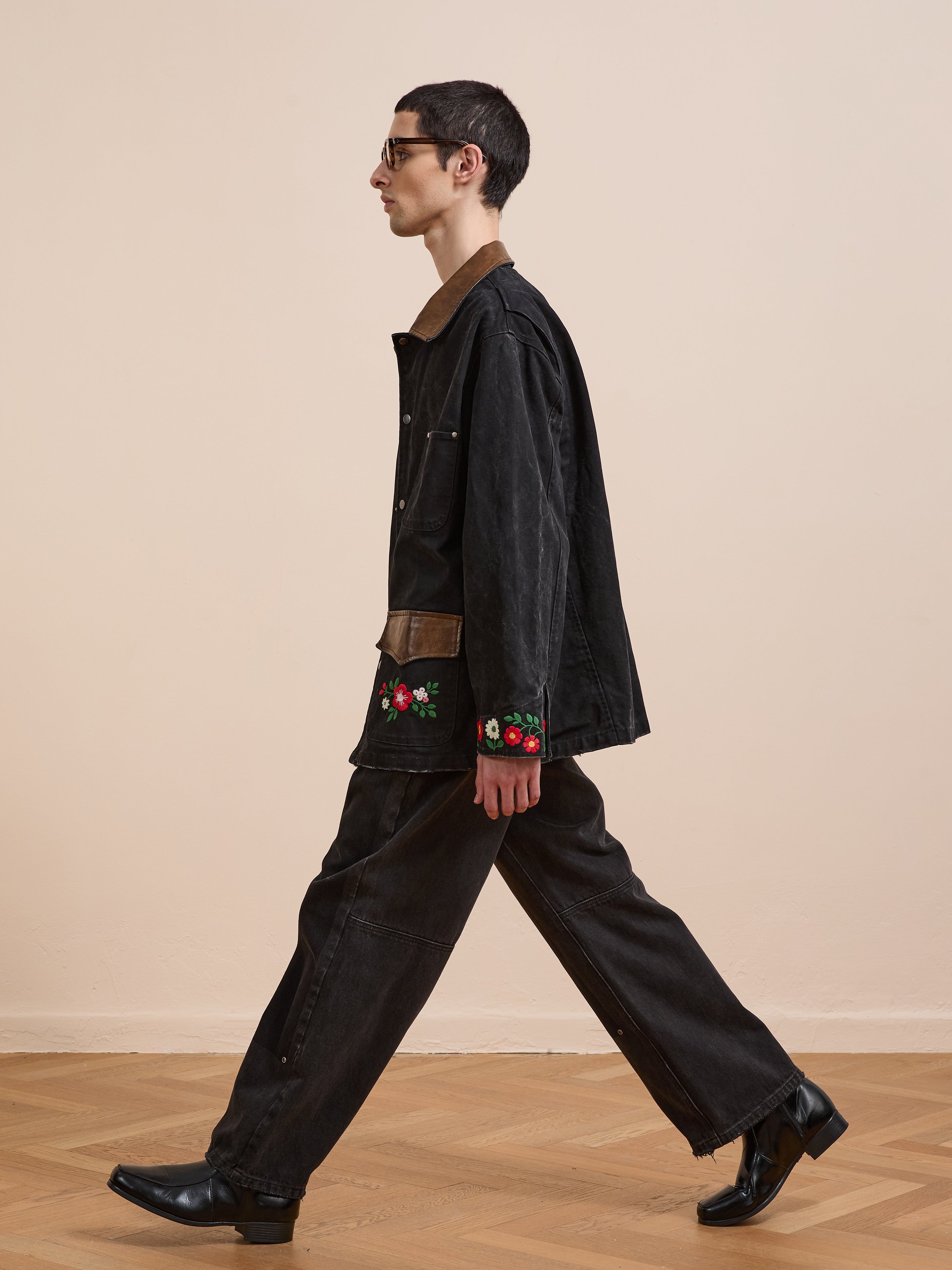 A person in sunglasses walks indoors wearing the Found Canvas Embroidered Work Jacket with matching pants, featuring floral embroidery, black shoes, a brown collar detail, and a unisex relaxed fit.
