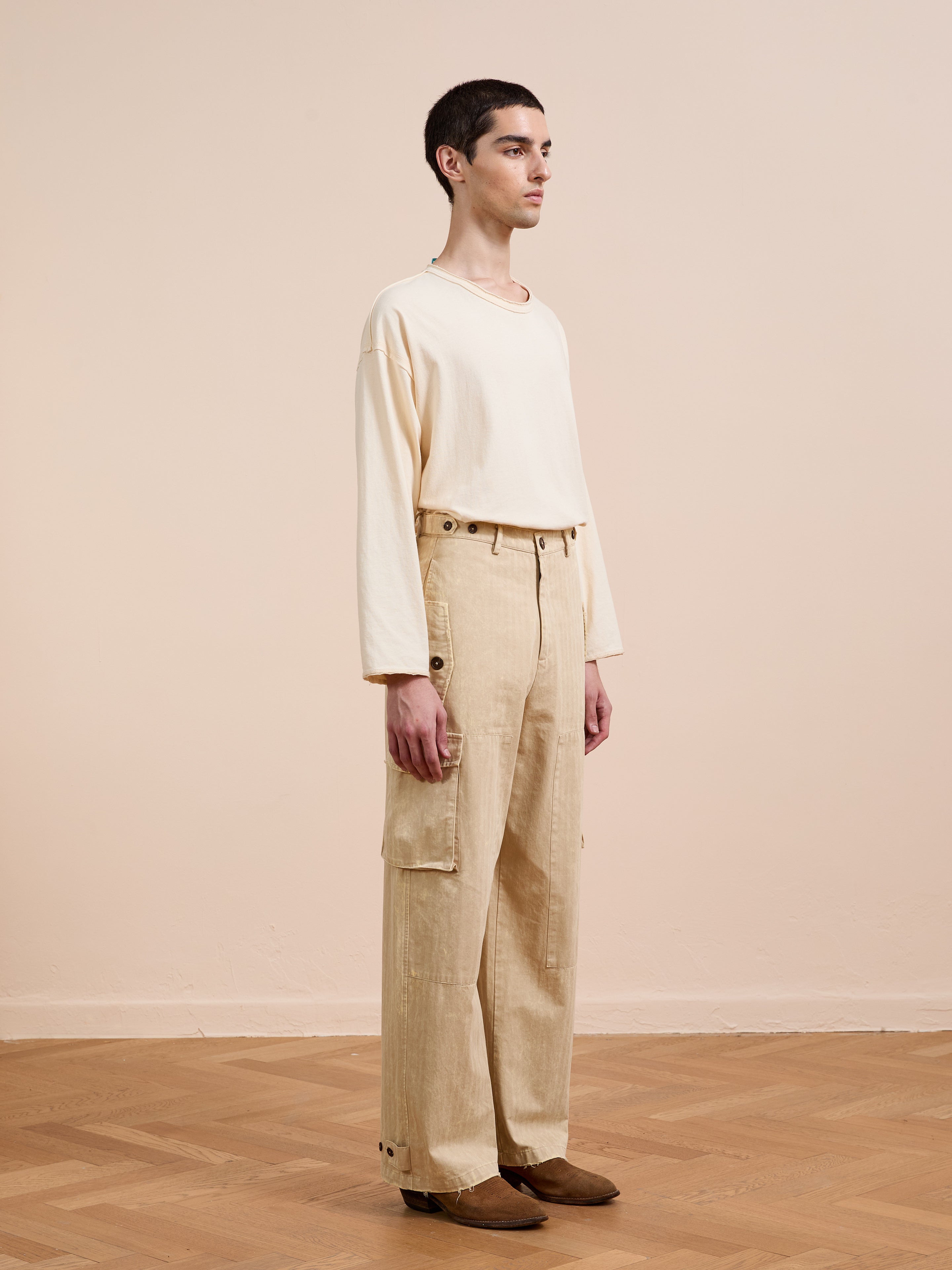A person stands indoors against a beige wall and wooden floor, wearing FOUND Utility Herringbone Paneled Pants with side buttons, a cream long-sleeve shirt, and brown shoes.
