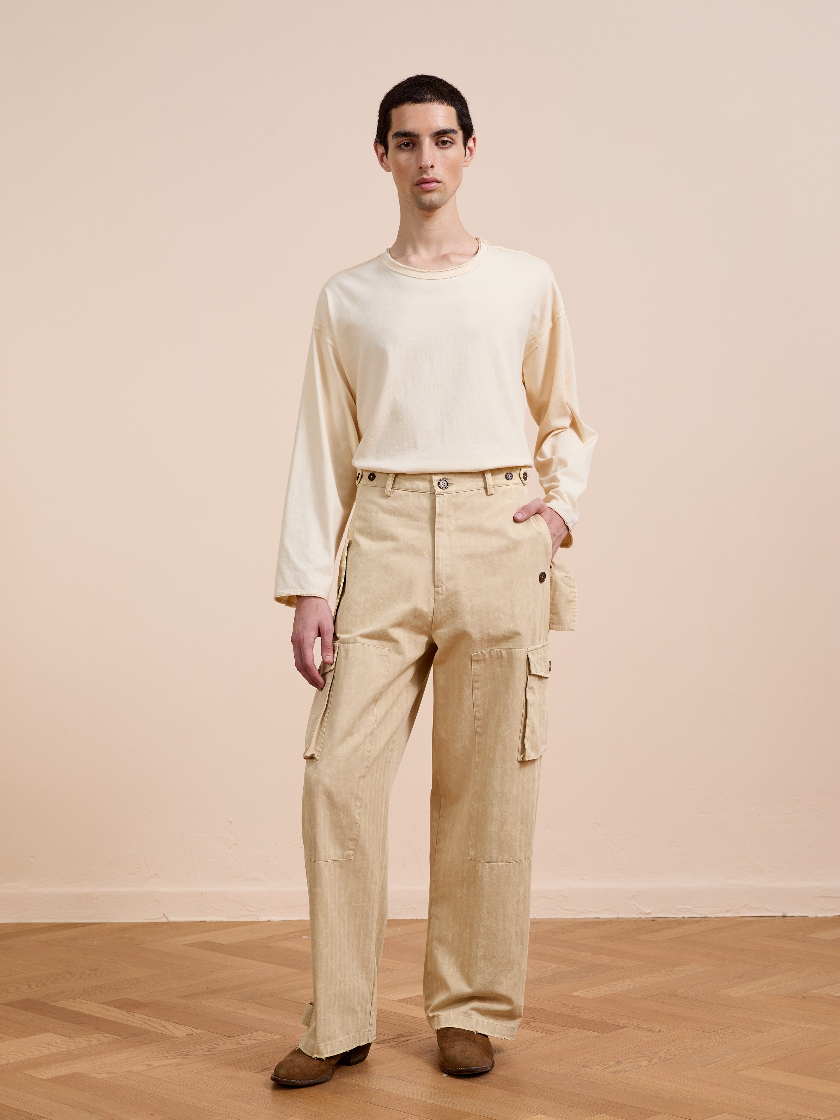 A person stands indoors against a plain light-colored wall and wooden floor, wearing FOUND’s Utility Herringbone Paneled Pants in beige with brown shoes and a cream long-sleeve shirt.