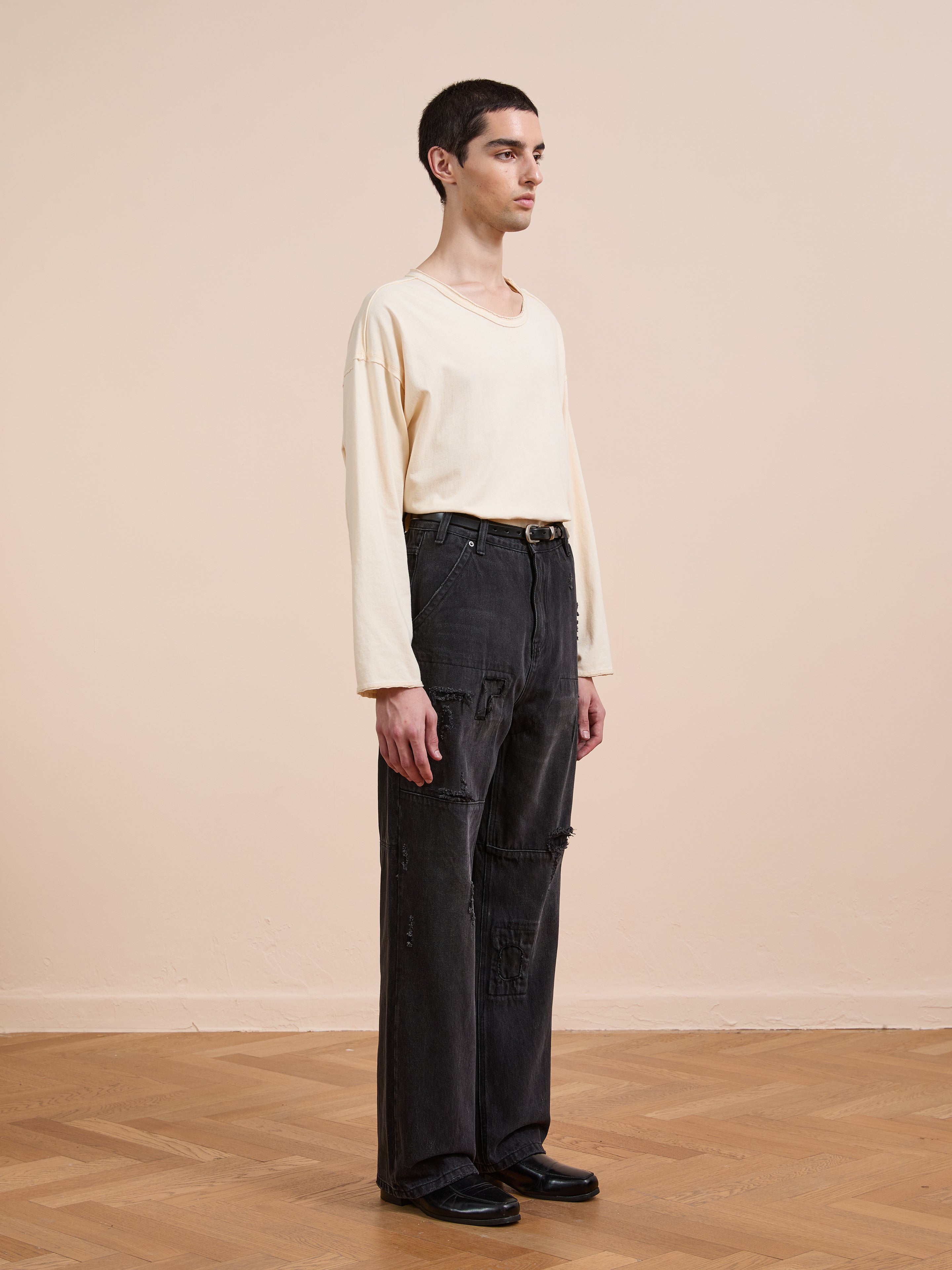 A person stands indoors on a wooden floor, facing left against a beige wall, wearing a cream long-sleeve shirt, black shoes, and FOUND Painter Repaired Jeans—a vintage wide-leg denim style.