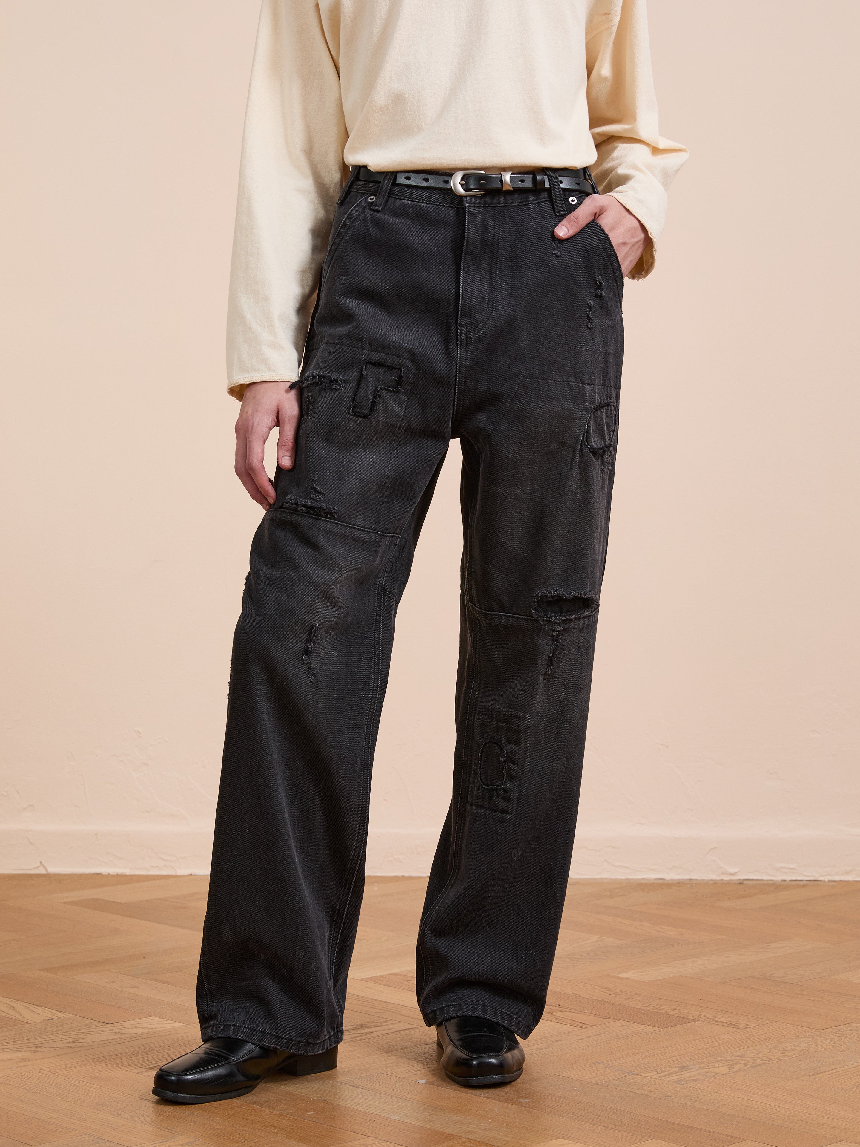 A person stands on a wooden floor against a beige wall, wearing a long-sleeve cream shirt, black belt, black shoes, and FOUND's Painter Repaired Jeans.