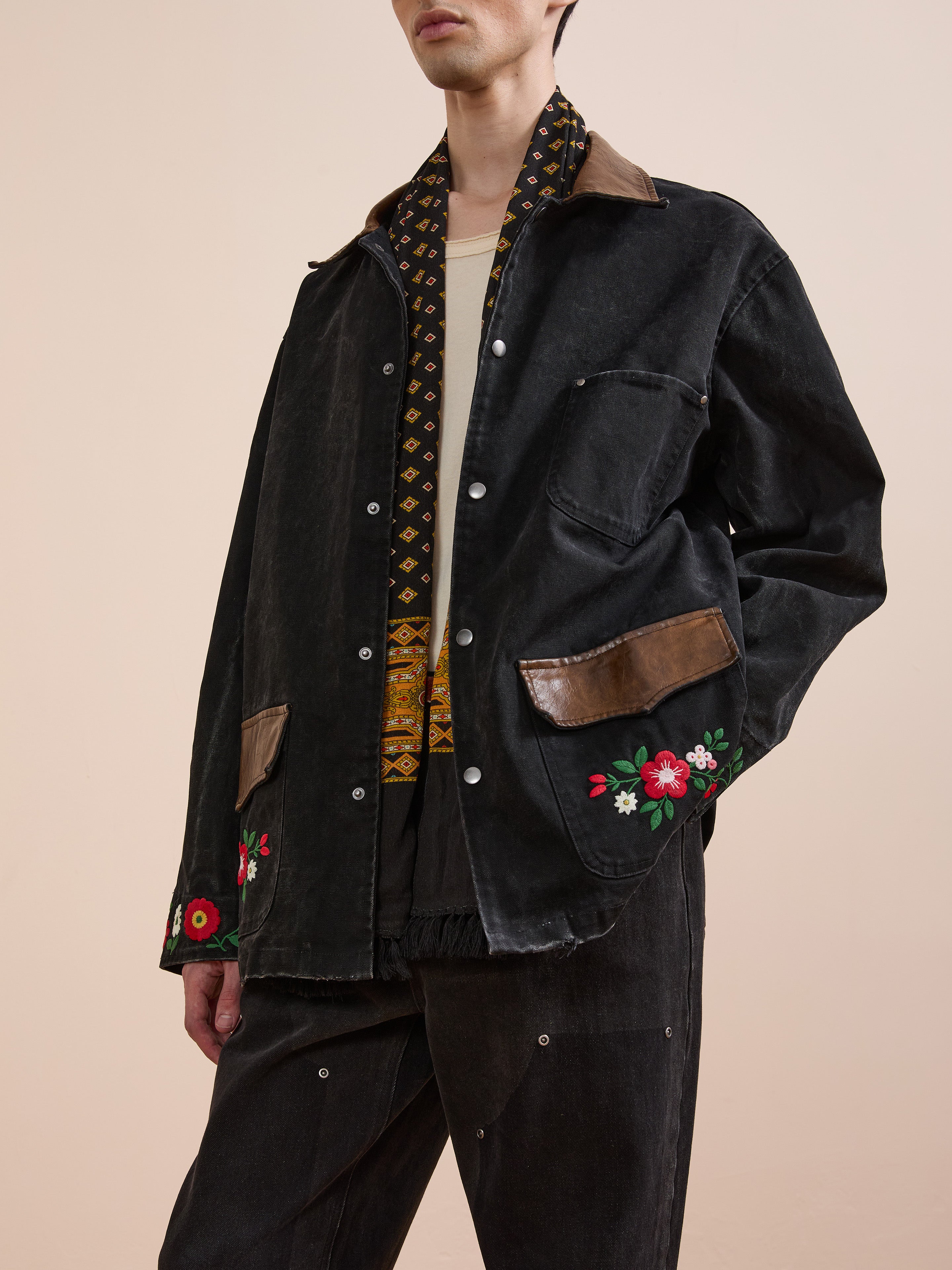 A person wearing Found’s Canvas Embroidered Work Jacket—black with brown accents and floral embroidery on the pockets—plus a light shirt and black pants, stands against a beige background. This unisex, relaxed-fit jacket offers comfort and style.
