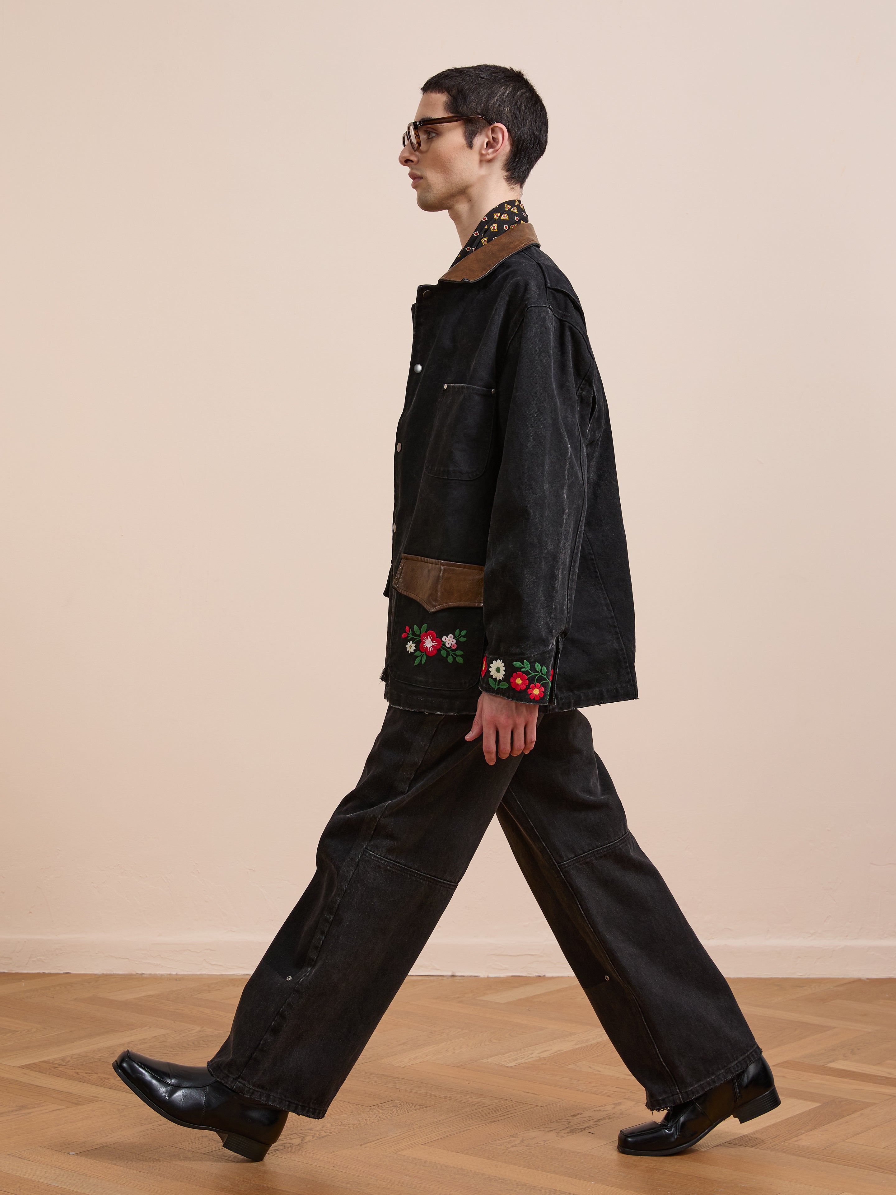 A person in Found's Canvas Embroidered Work Jacket with floral sleeve details and black shoes walks across a wooden floor against a plain beige wall.