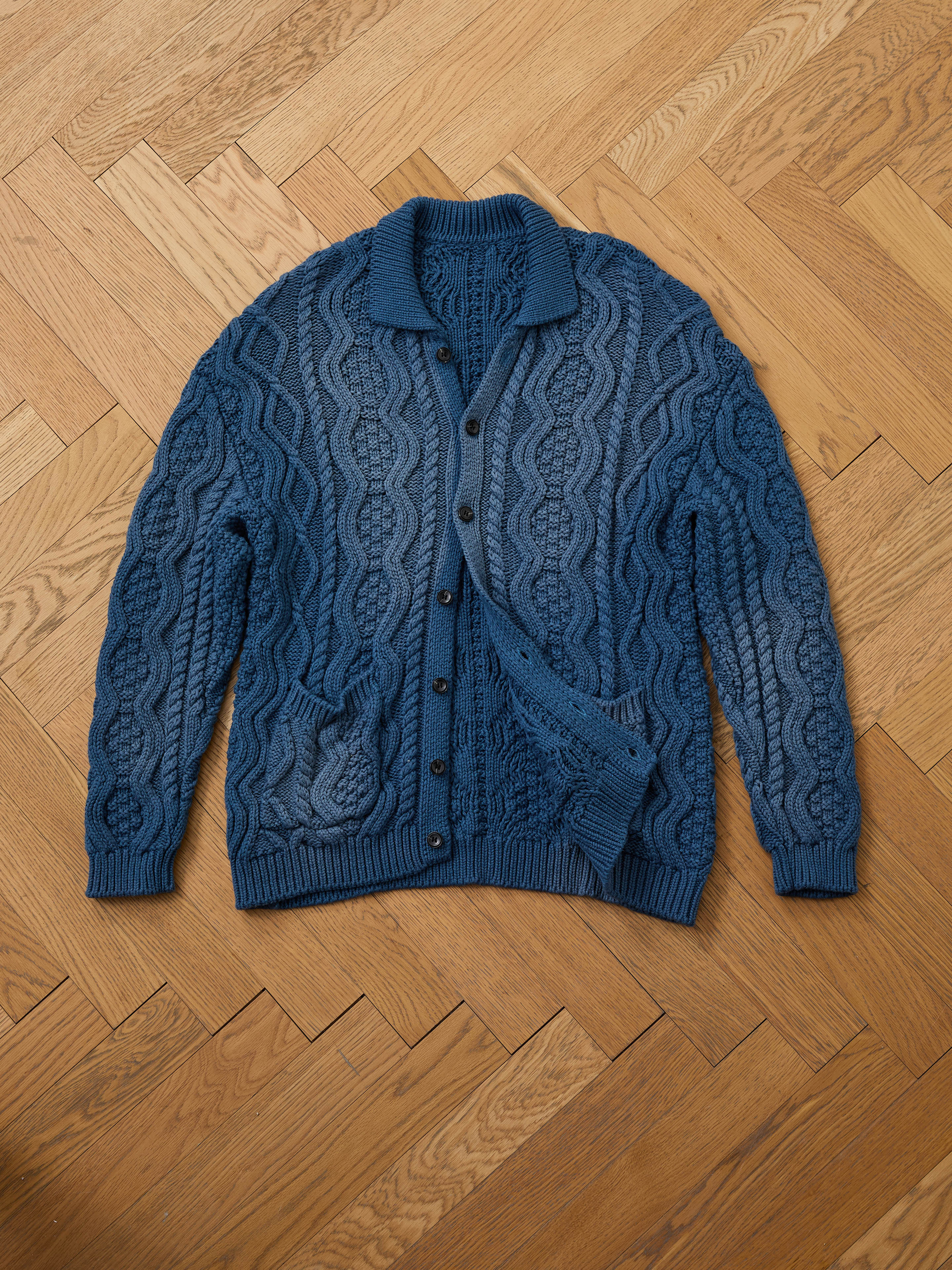 The Found Cable Knit Buttoned Cardigan in navy is displayed flat on a wooden herringbone-patterned floor.