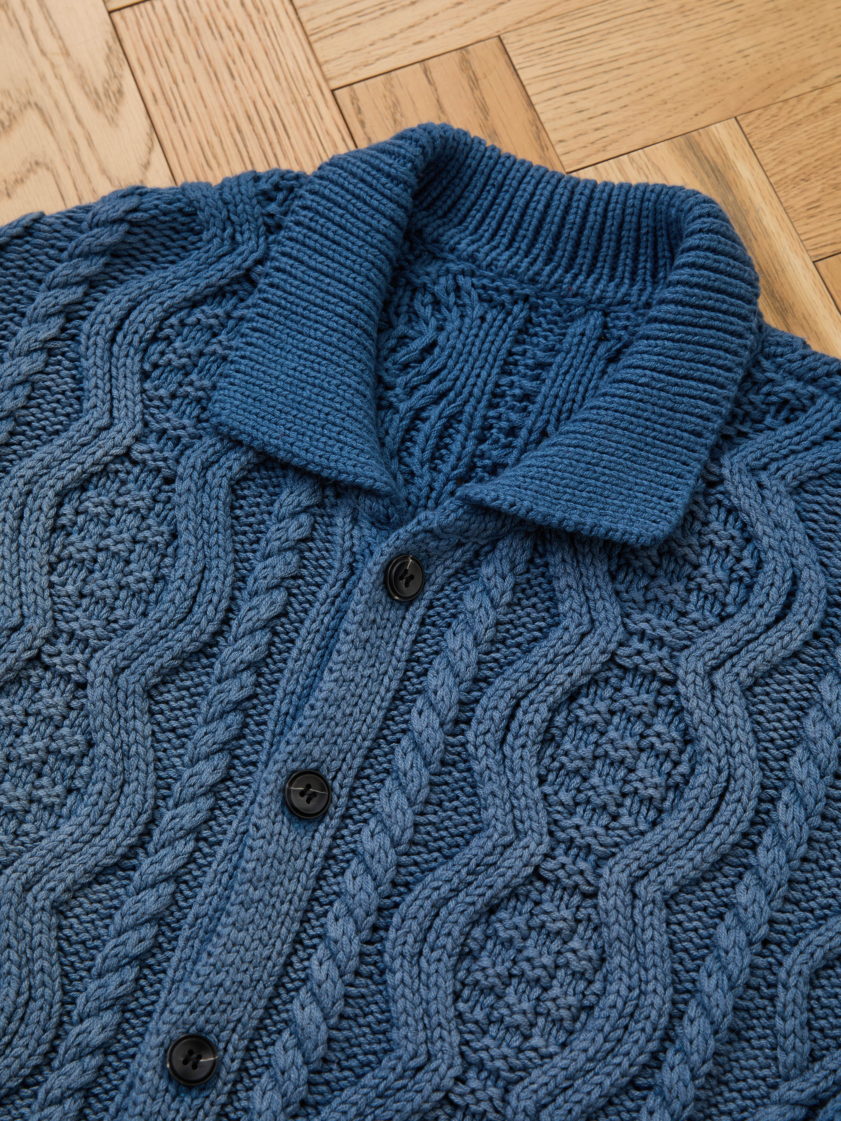 A close-up of the Found Cable Knit Buttoned Cardigan, featuring a textured navy knit, ribbed collar, and black buttons, displayed on a wooden parquet floor.
