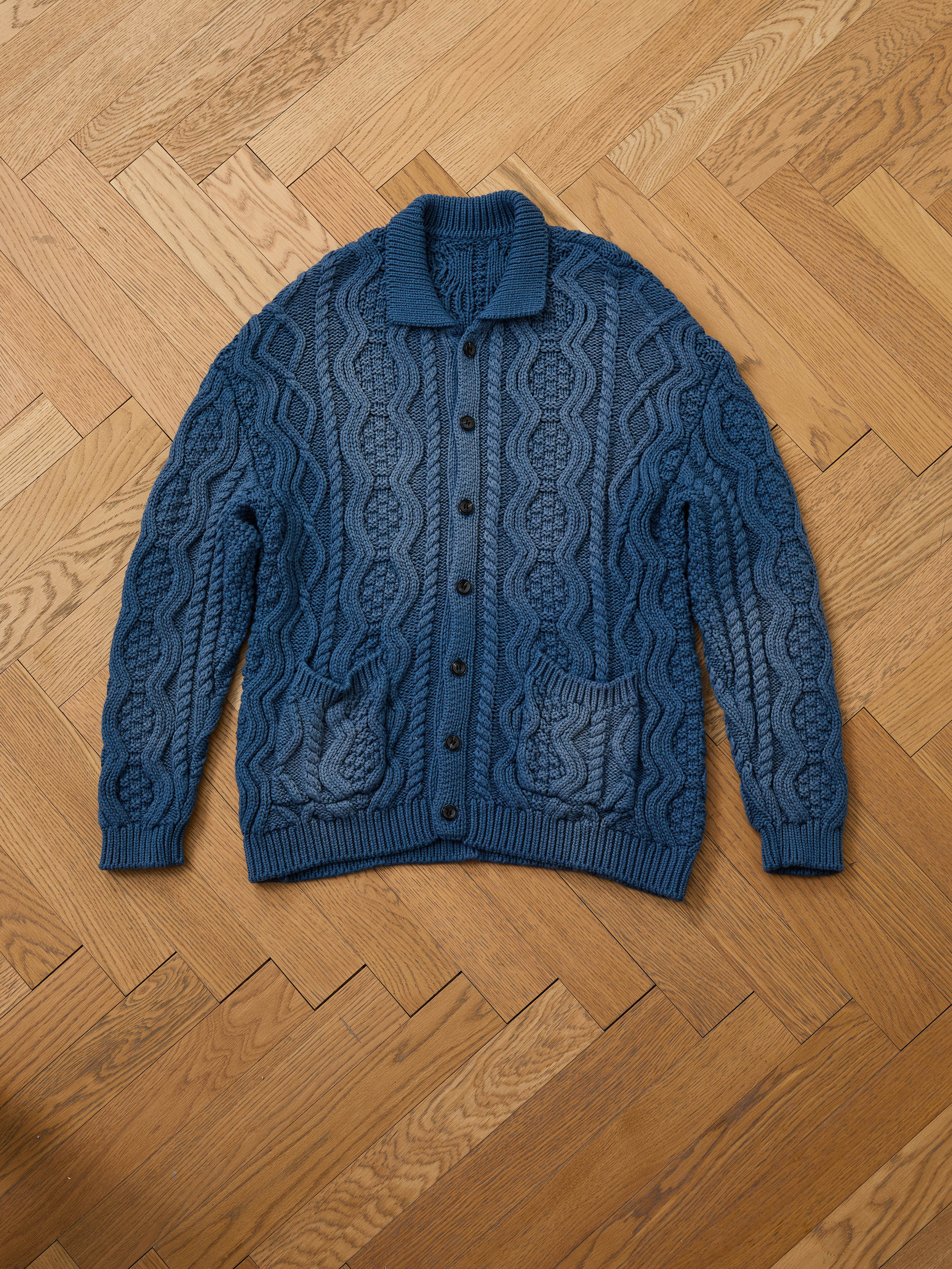 The Found Cable Knit Buttoned Cardigan in navy, featuring black buttons and two front pockets, is laid flat on a light brown herringbone wood floor, highlighting its classic unisex design.