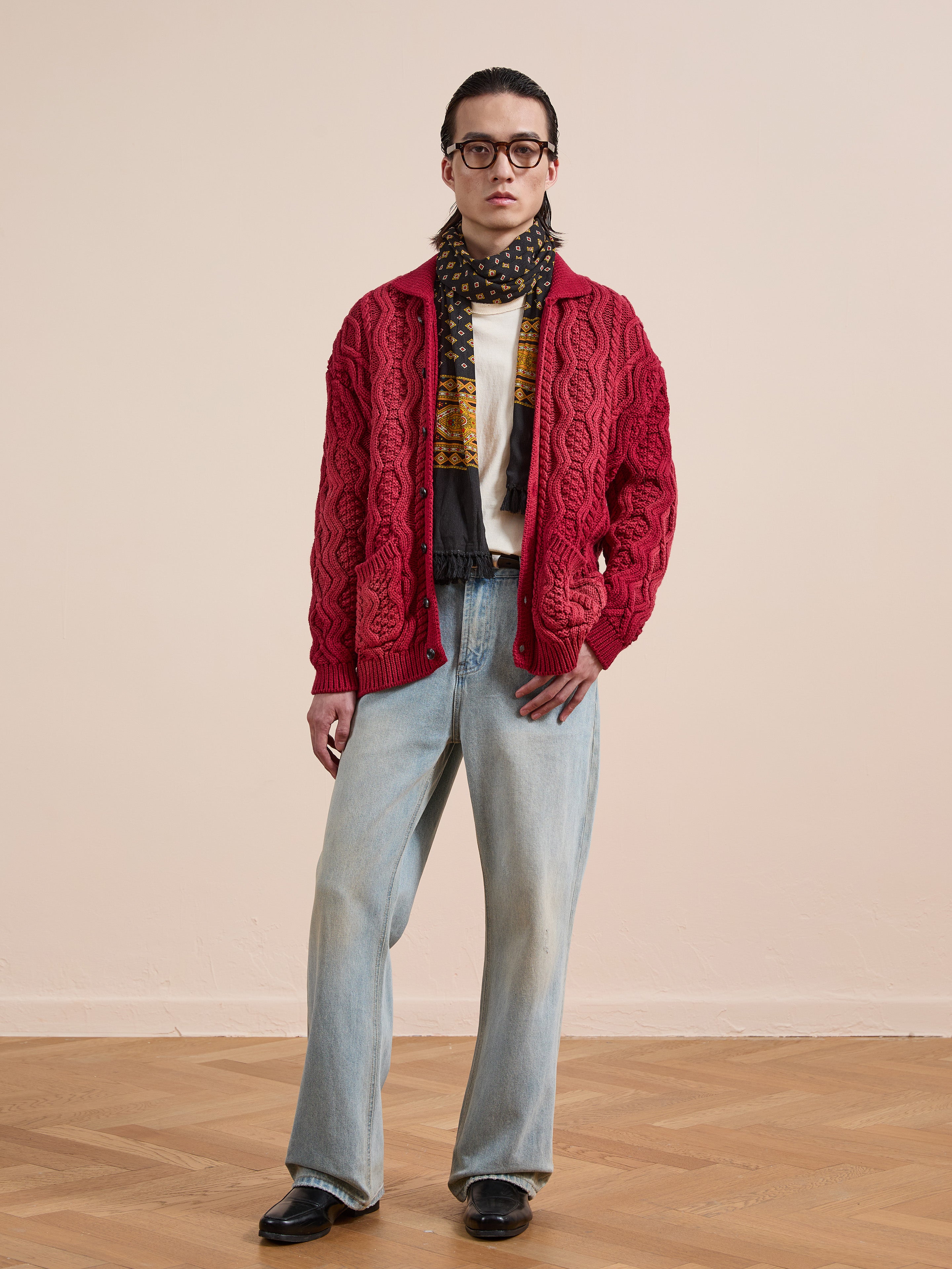 A person wears the FOUND Cable Knit Buttoned Cardigan in red, with a patterned scarf, white tee, light blue wide-leg jeans, and black shoes, standing on a wooden floor against a plain wall.