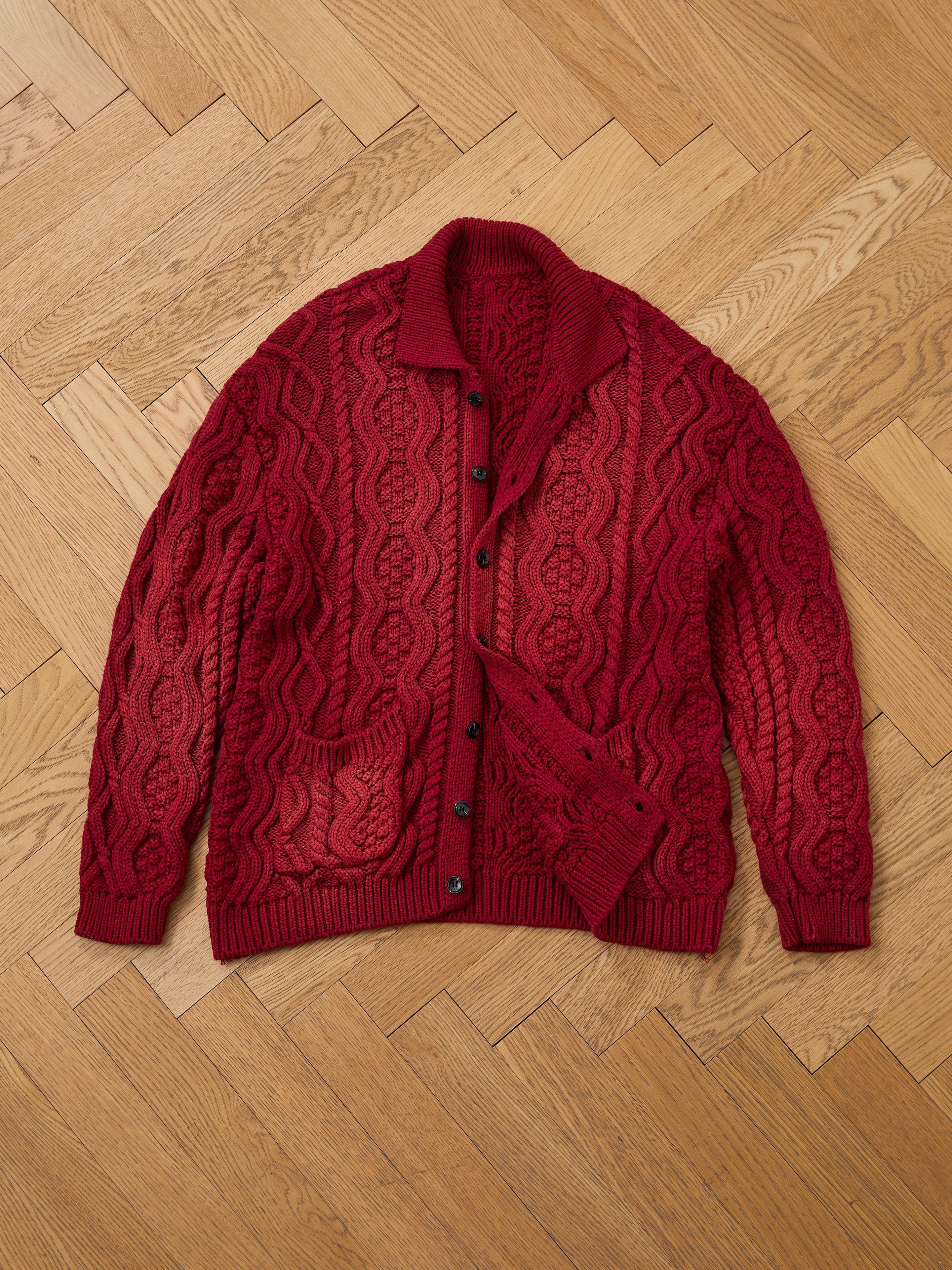 The FOUND Cable Knit Buttoned Cardigan in red, featuring front pockets, is laid flat on a herringbone-patterned wooden floor.