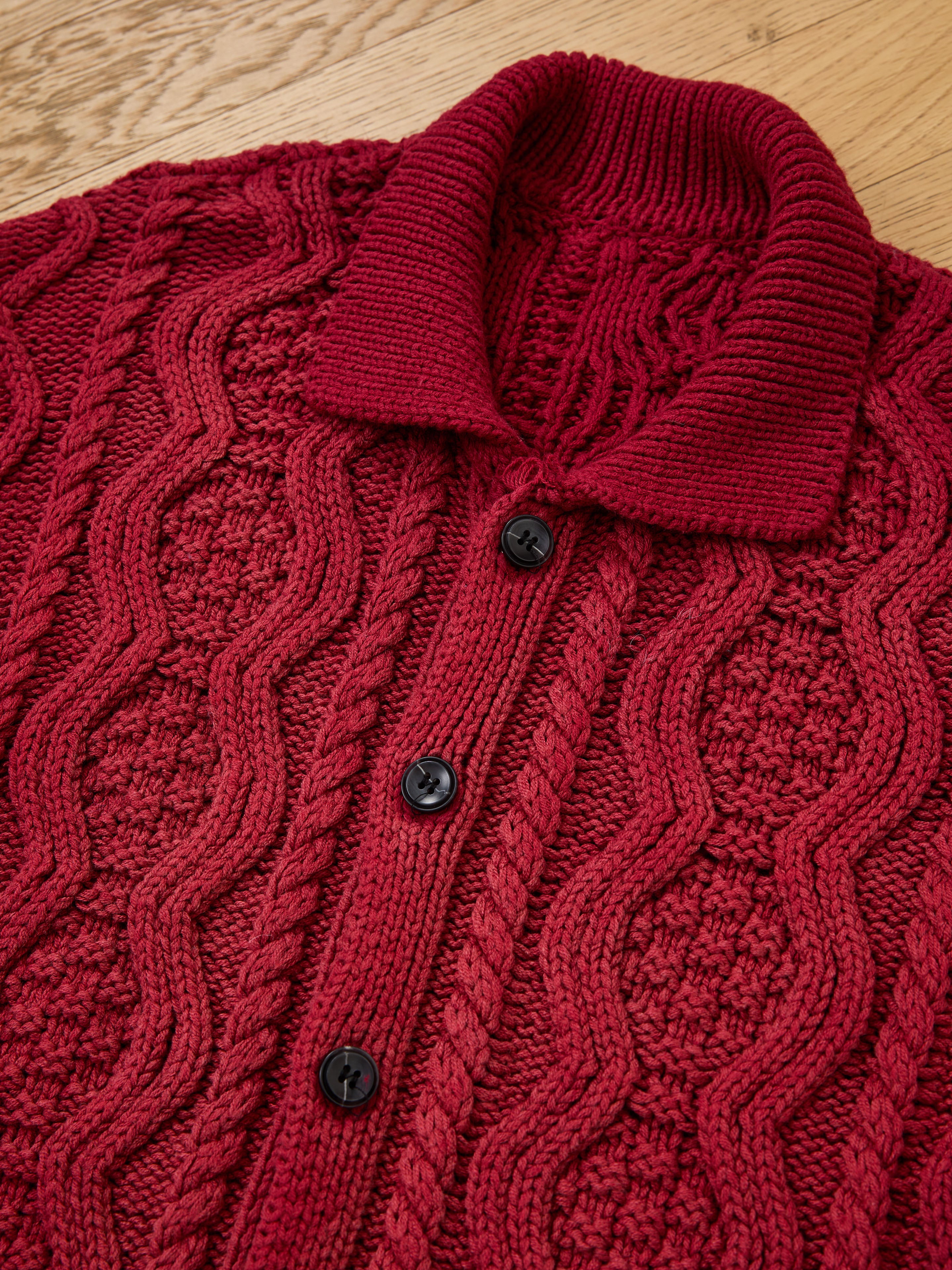 A close-up of the FOUND Cable Knit Buttoned Cardigan in red, featuring black buttons and a vintage-dyed finish, highlights its textured pattern as it lays flat on a wooden surface.