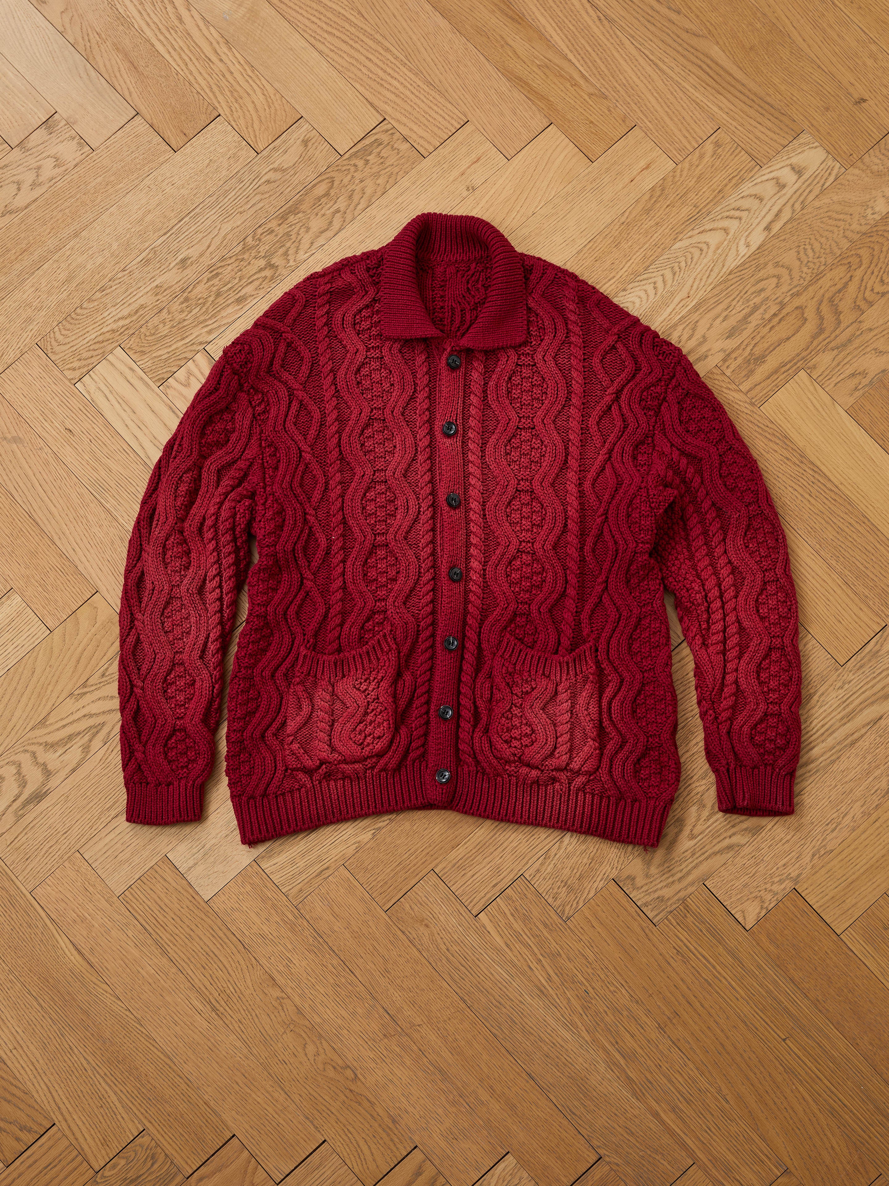 The FOUND Cable Knit Buttoned Cardigan in red, featuring two front pockets and a chunky knit, is displayed flat on a wooden herringbone floor.