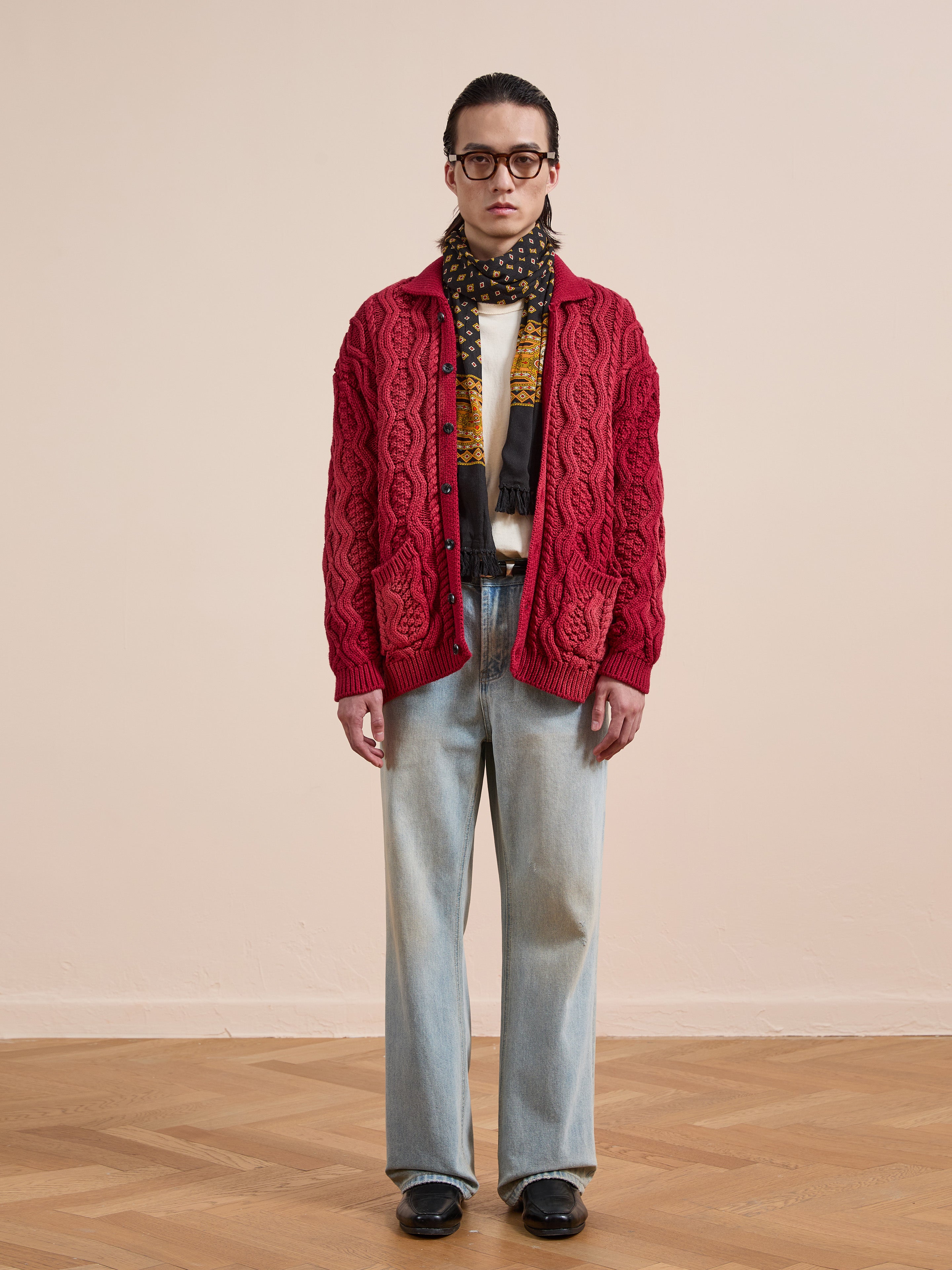 A person wears the FOUND Cable Knit Buttoned Cardigan in red, light blue jeans, a polka dot scarf, glasses, and black shoes while standing indoors on a wooden floor against a plain beige wall.