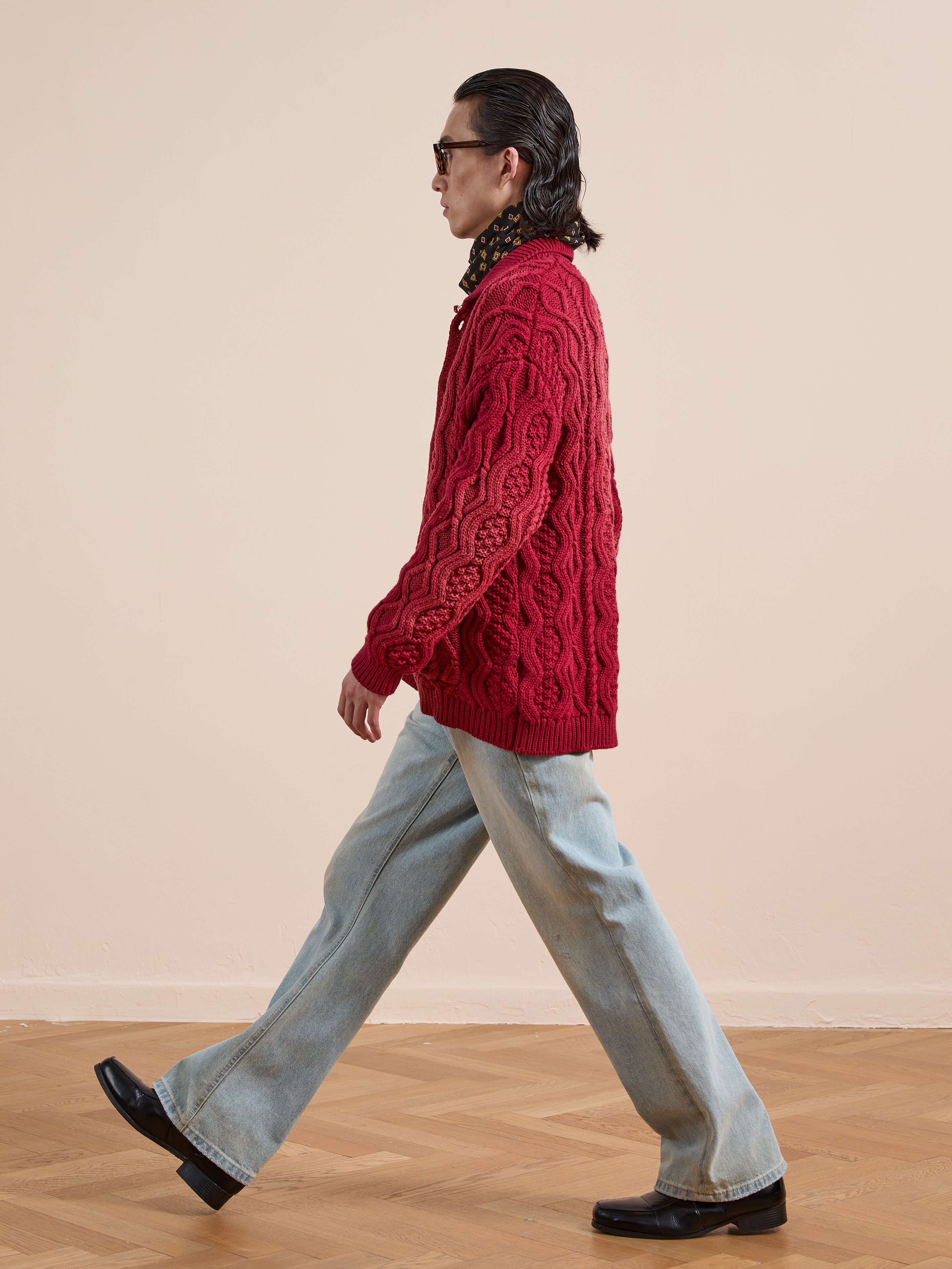 Wearing sunglasses and the FOUND Cable Knit Buttoned Cardigan in red, along with wide-leg light blue jeans and black shoes, a person walks indoors on a wooden floor.