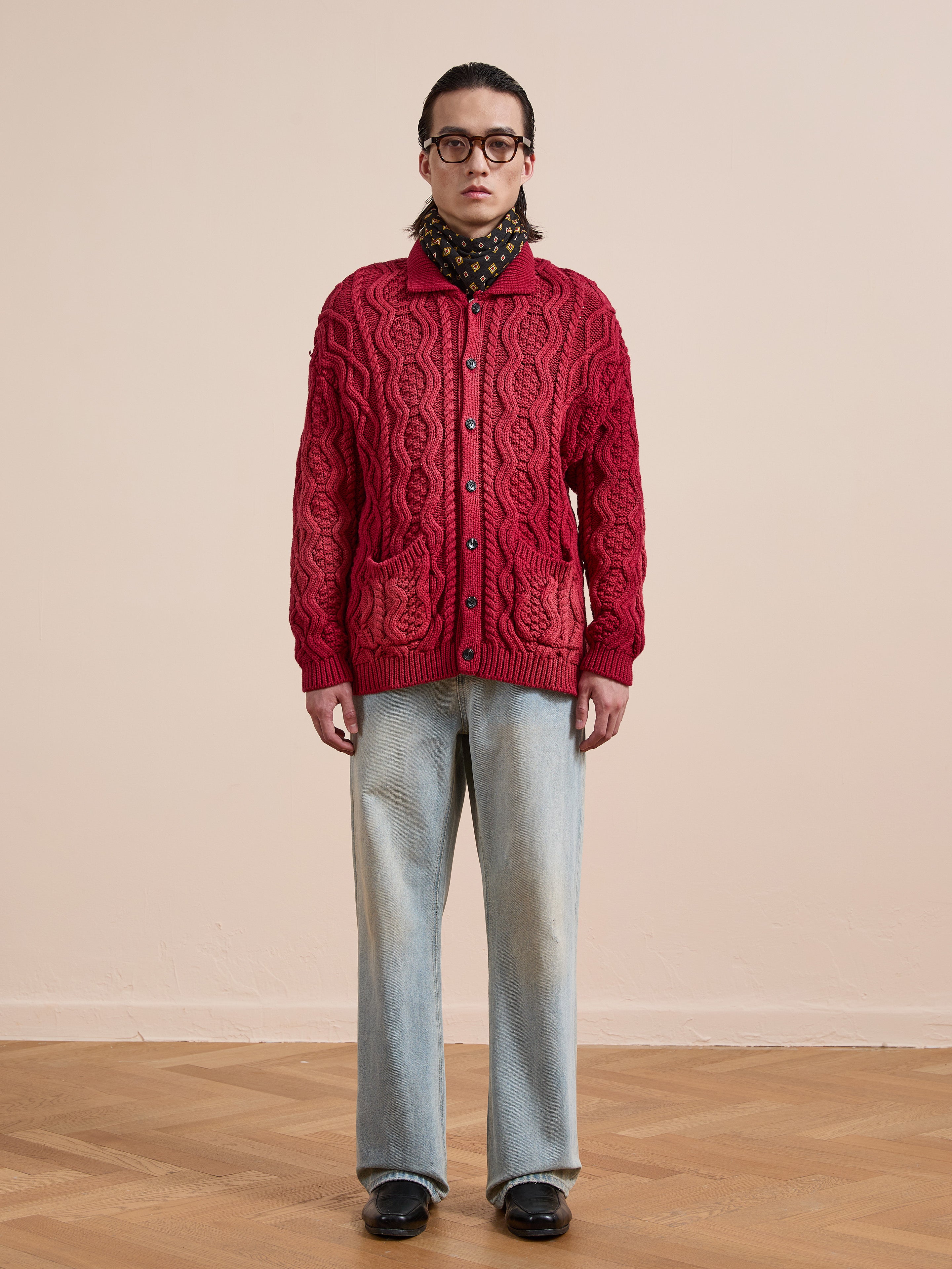 Person indoors against a beige wall, wearing the FOUND Cable Knit Buttoned Cardigan in red, light blue jeans, black shoes, glasses, and a patterned scarf.