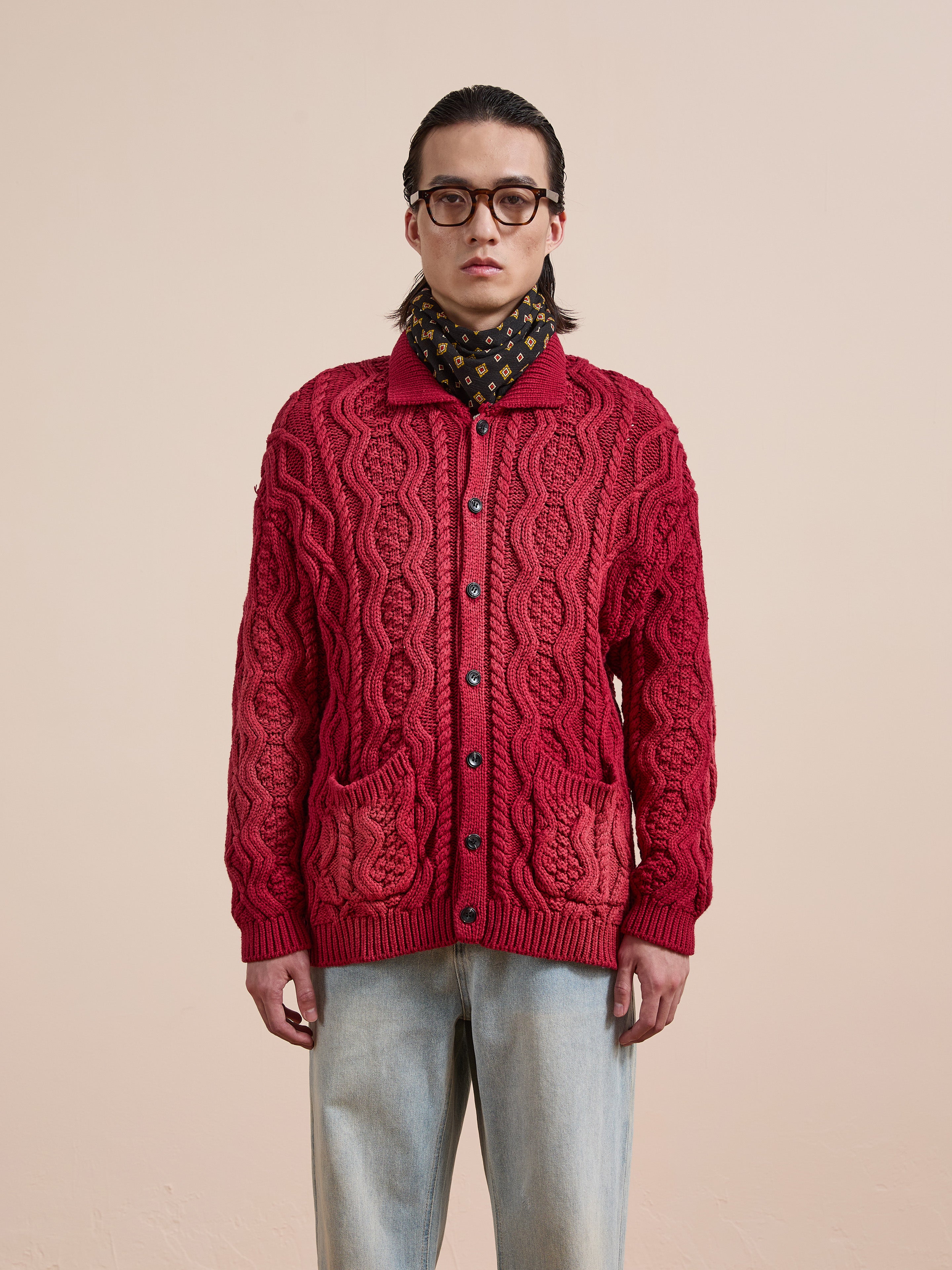 A person wearing glasses, a patterned scarf, light blue jeans, and the FOUND Cable Knit Buttoned Cardigan with front pockets stands against a plain background.