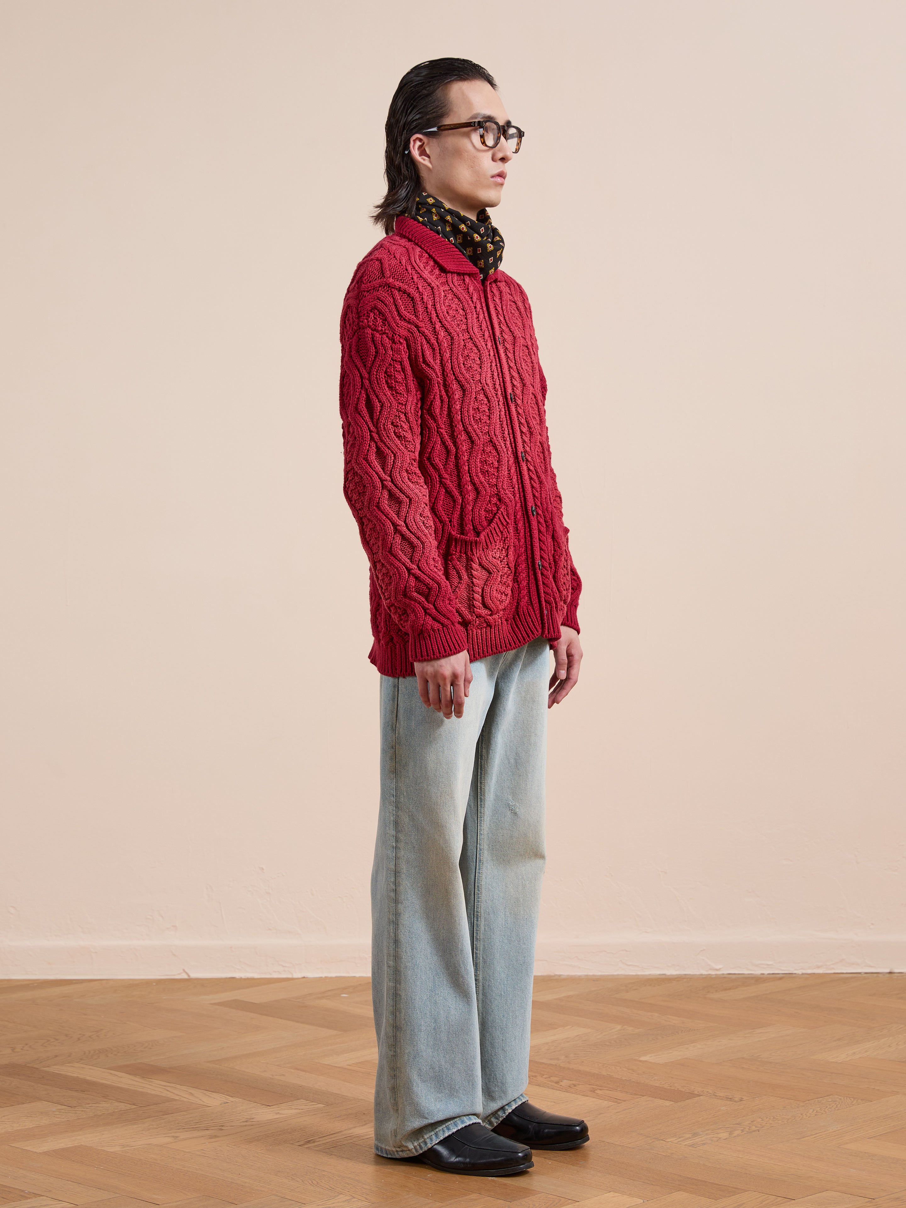 A person in profile wears the FOUND Cable Knit Buttoned Cardigan in red, paired with wide-leg light blue jeans, black shoes, and glasses. They stand on a wooden floor against a plain beige background.