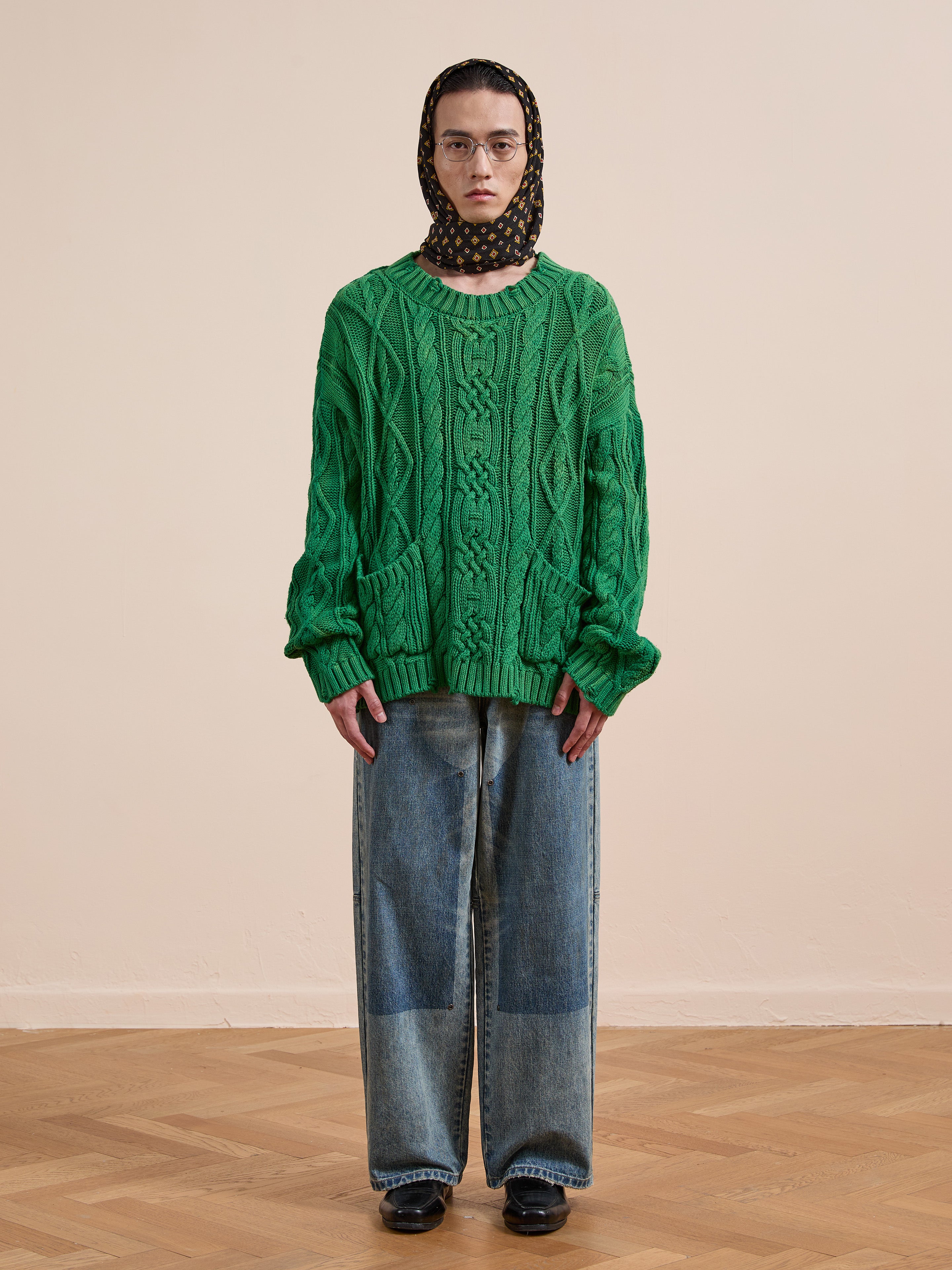 A person stands indoors wearing the Found Cable Knit Pullover Sweater in green, paired with loose blue jeans, black shoes, glasses, and a polka dot headscarf against a plain beige background.