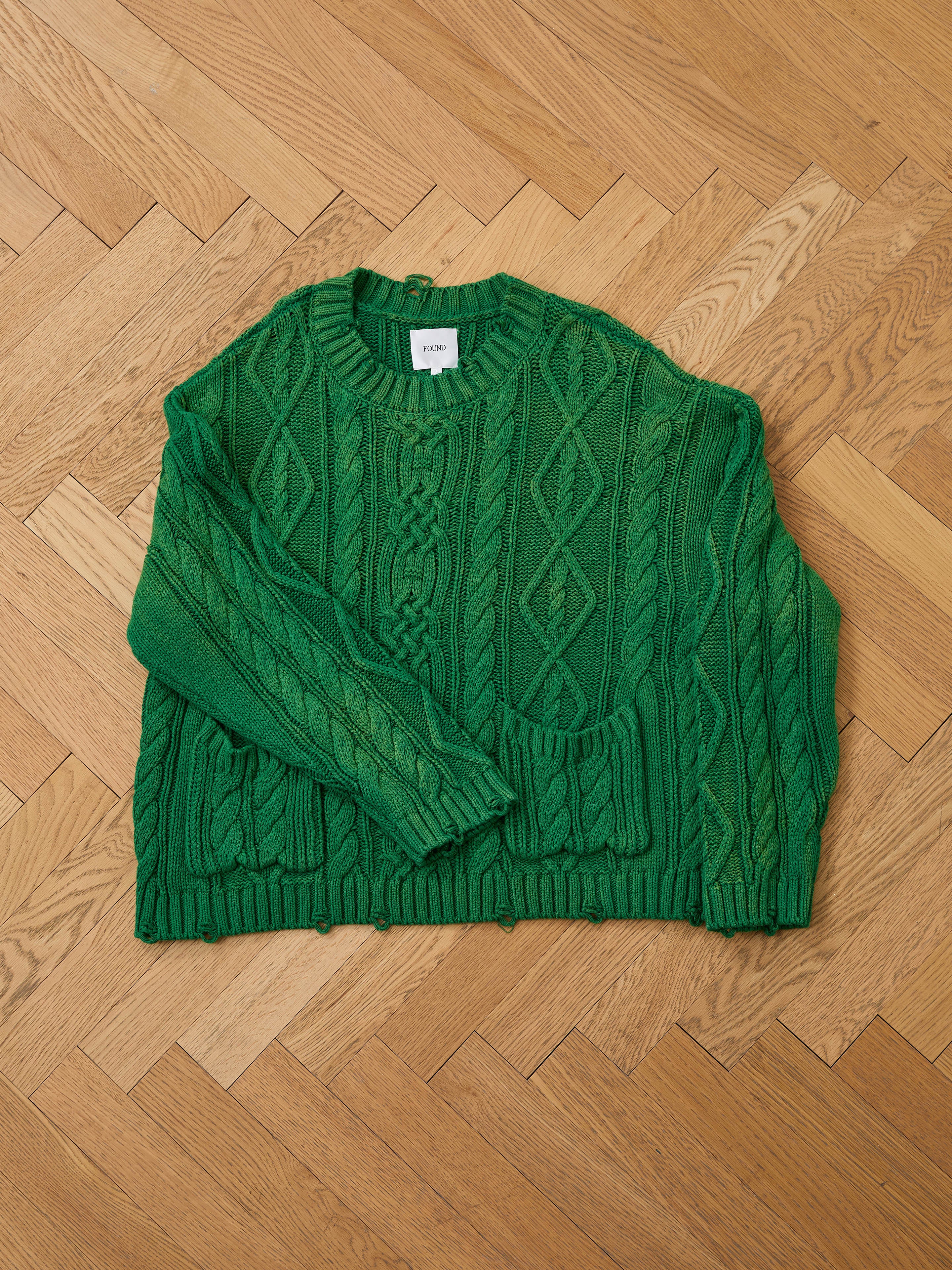 The Found Cable Knit Pullover Sweater is a cozy, unisex green knit with long sleeves, shown laid flat on a light wooden herringbone floor.
