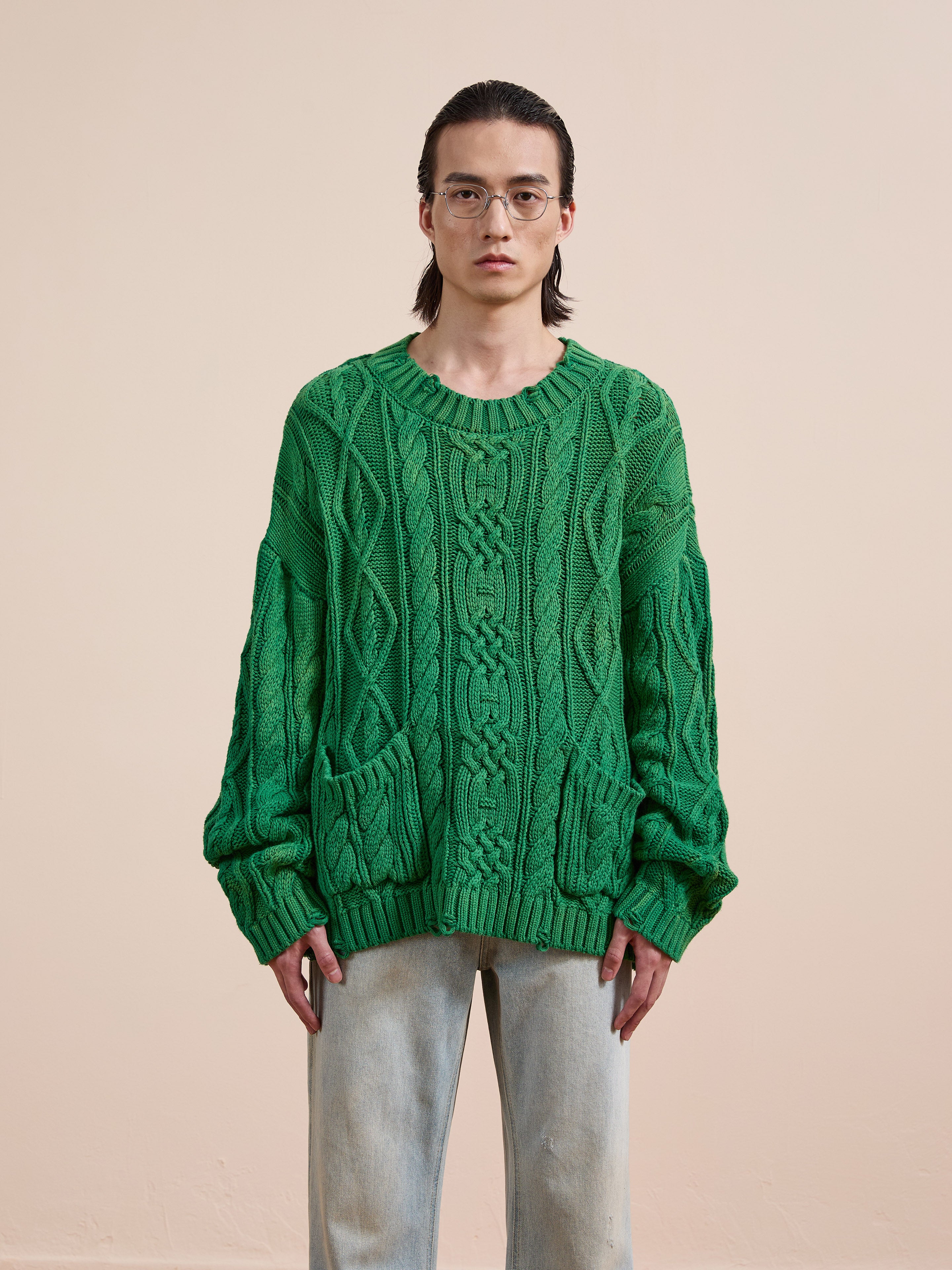 A person stands against a light beige background wearing the Found Cable Knit Pullover Sweater in green, featuring front pockets, paired with light blue jeans.