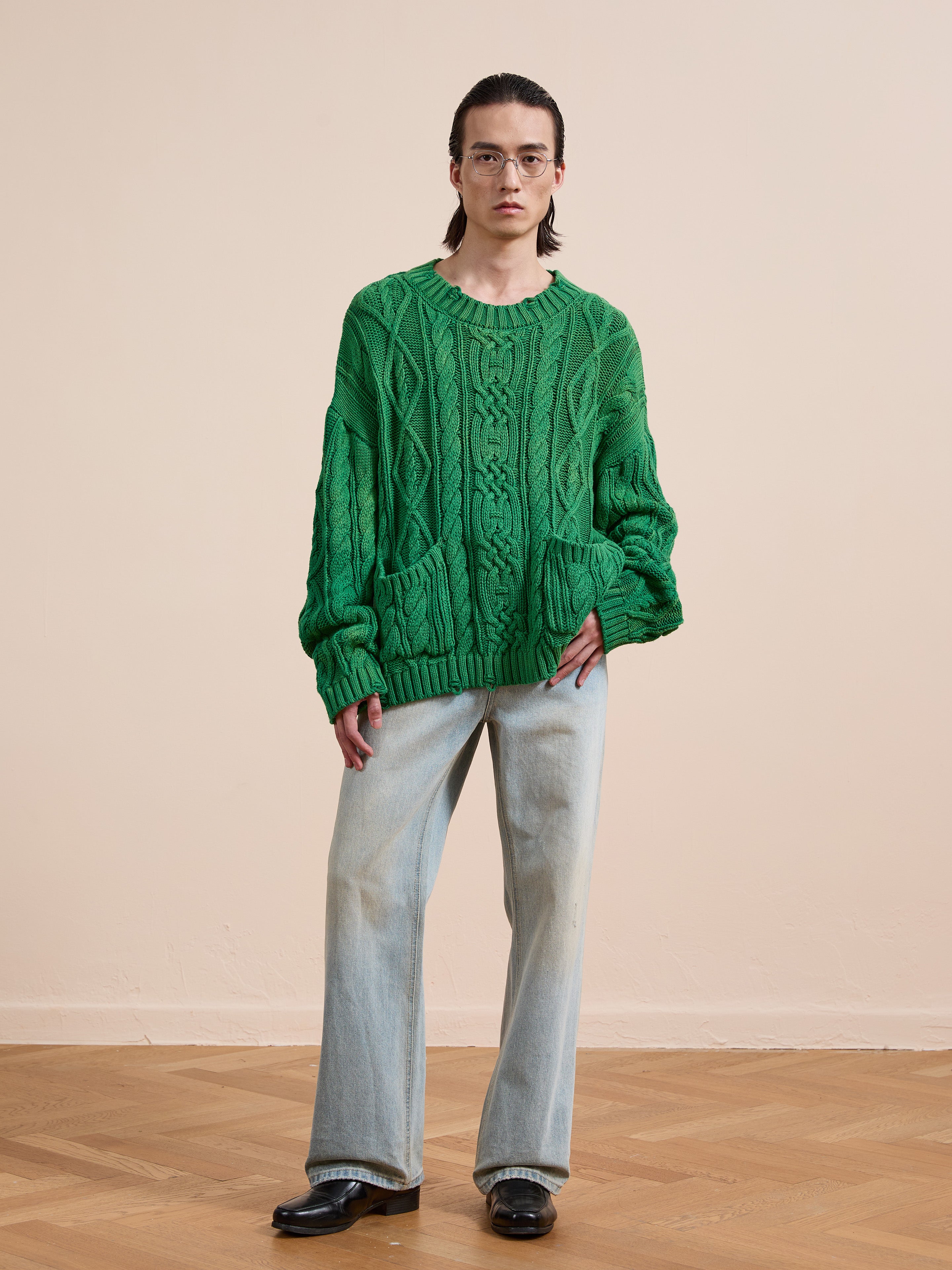 Indoors, a person wears Found's Cable Knit Pullover Sweater, glasses, light blue wide-leg jeans, and black shoes on a wooden floor with a plain beige wall background.