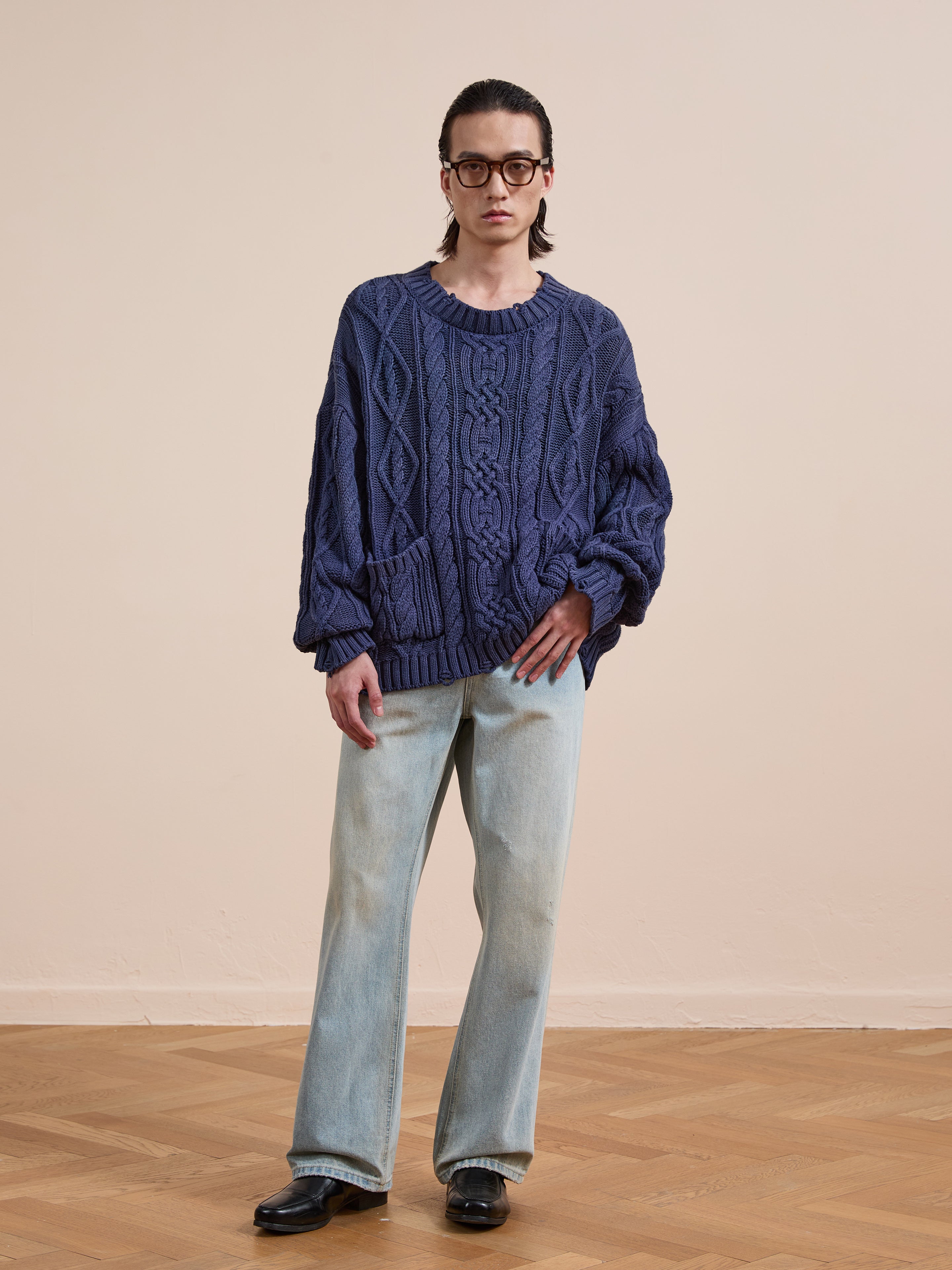 A person wears the Found Cable Knit Pullover Sweater in blue, paired with light blue jeans and black shoes, standing indoors on a wooden floor against a beige wall.