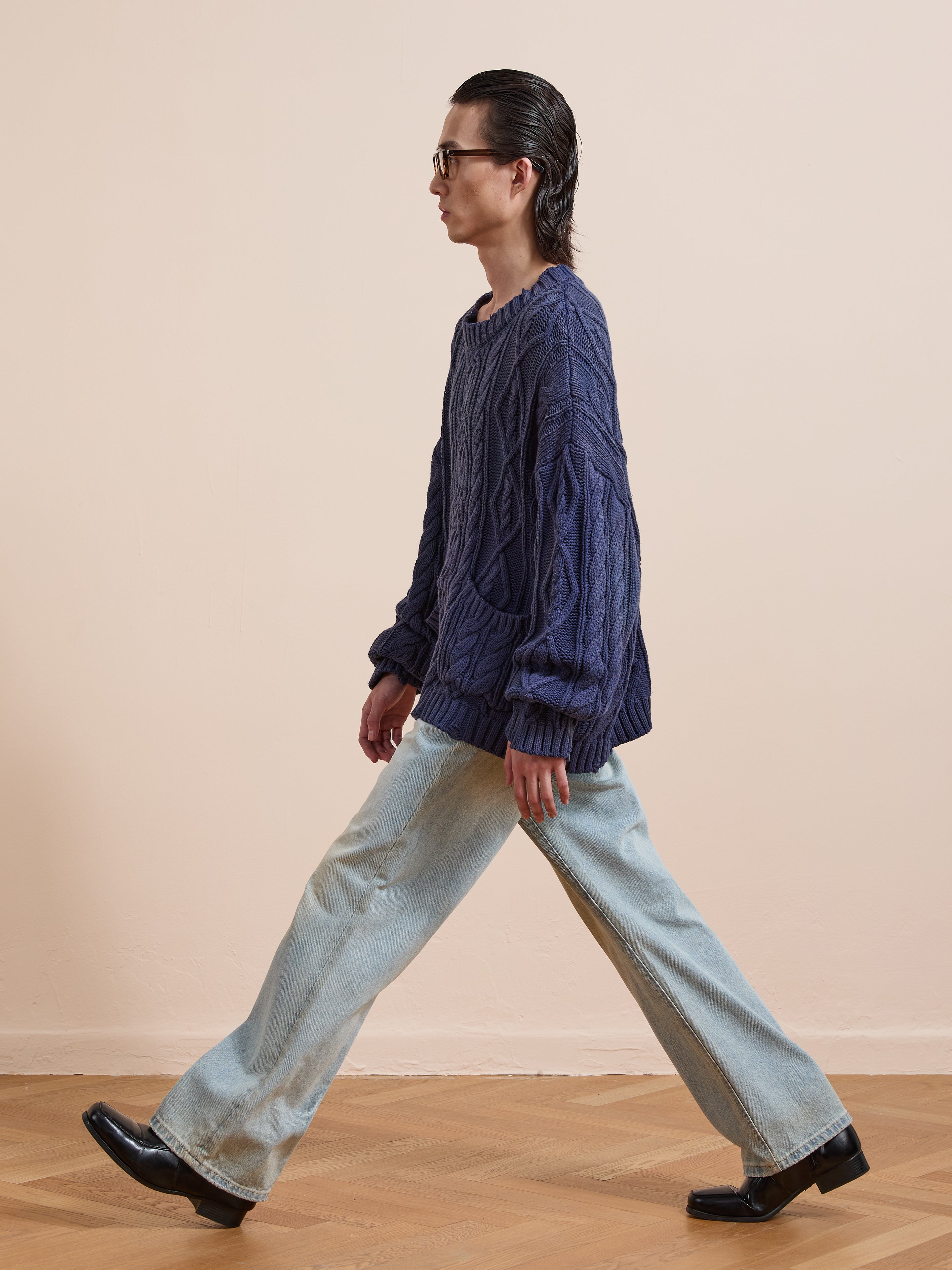 A person with long dark hair and glasses wears the Found Cable Knit Pullover Sweater in navy, light blue wide-leg jeans, and black shoes, walking indoors on a wooden floor against a beige wall for a cozy unisex style.