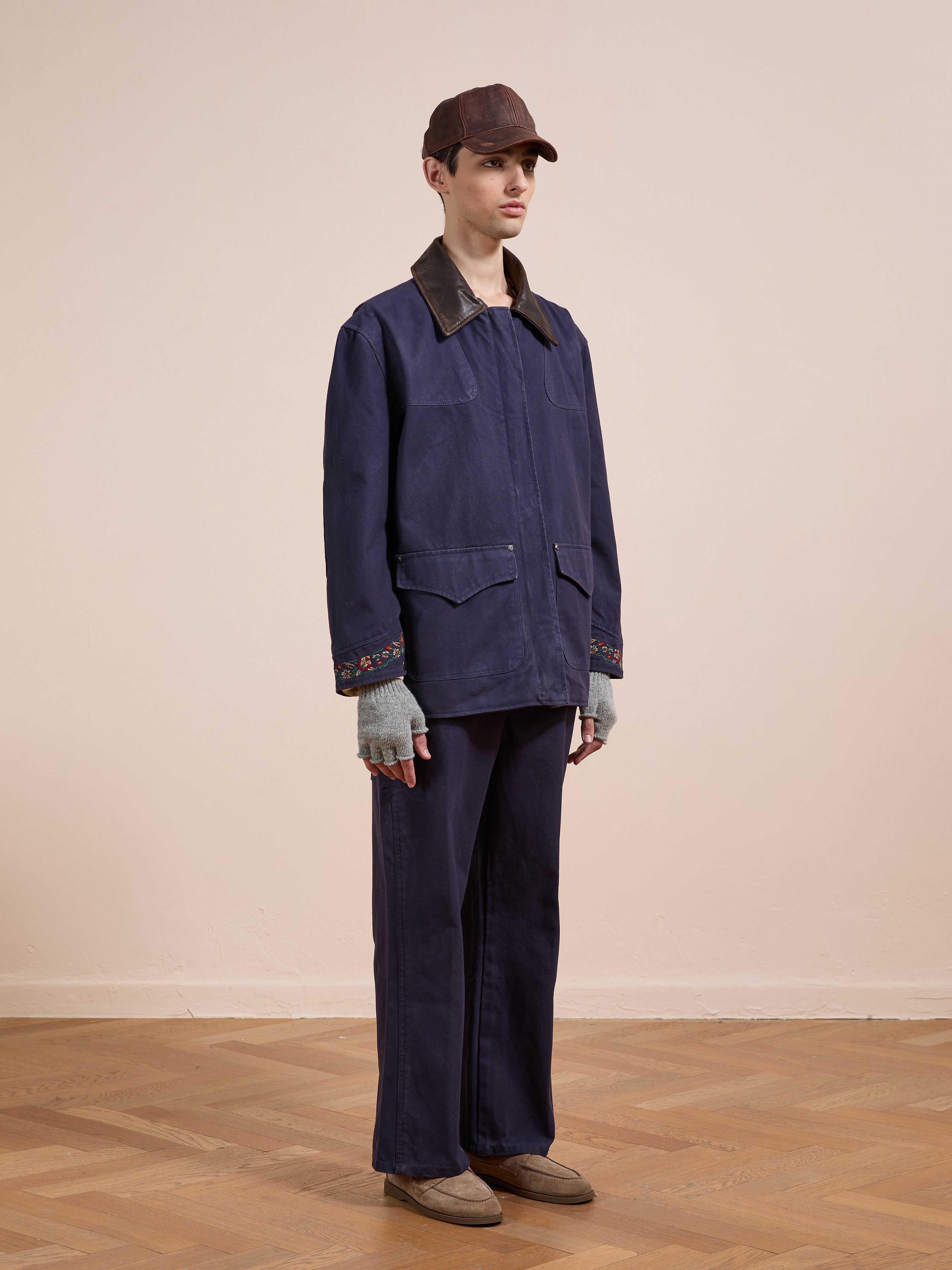 A person indoors wears the Found Canvas Distressed Barn Jacket with a leather collar, matching pants, brown cap, gray fingerless gloves, and brown shoes. The unisex, relaxed fit stands out against a plain background and wooden floor.