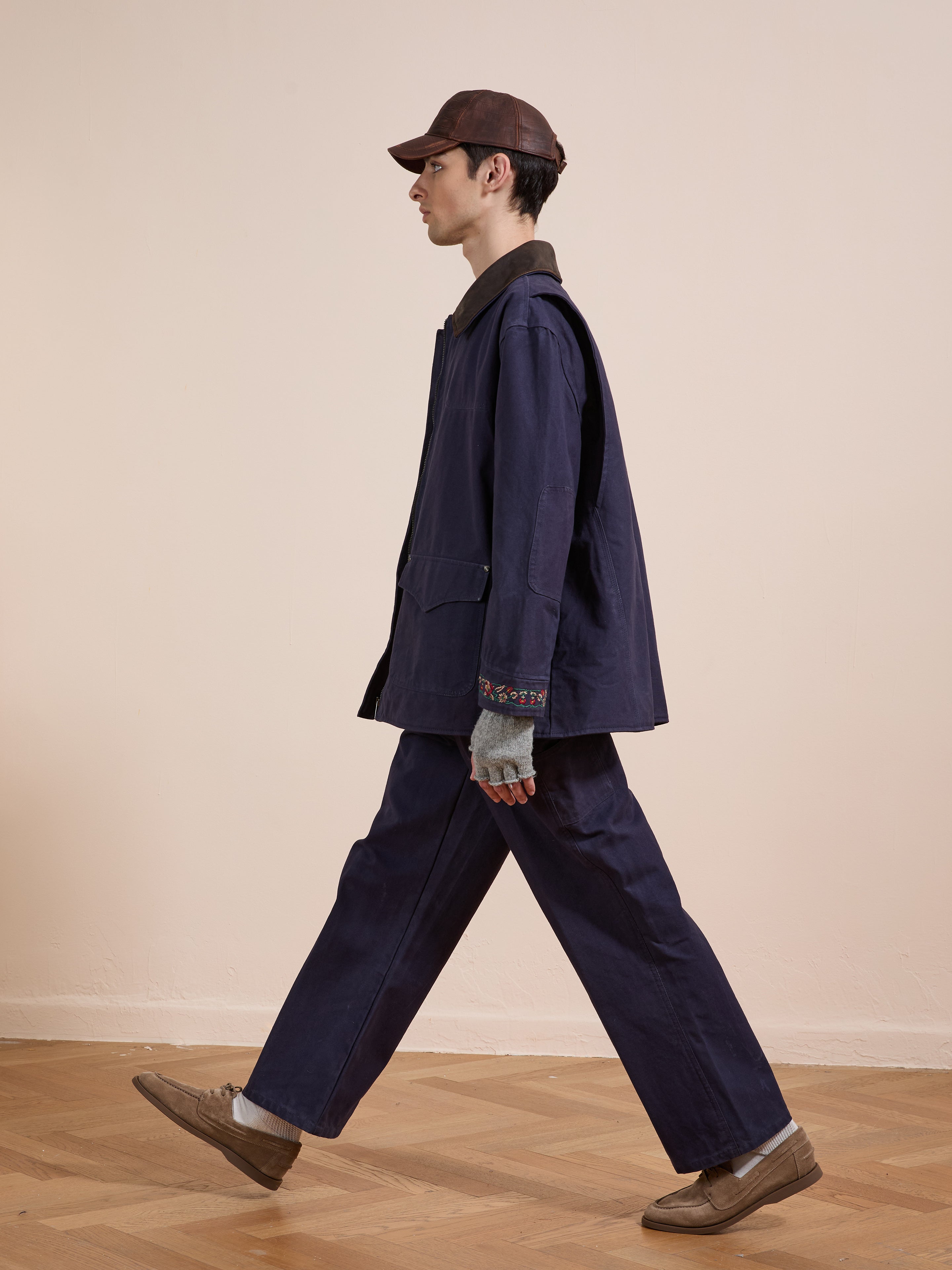 A person wearing the Found Canvas Distressed Barn Jacket with a leather collar, gray gloves, brown shoes, and a brown cap walks indoors on a wooden floor against a plain beige wall.