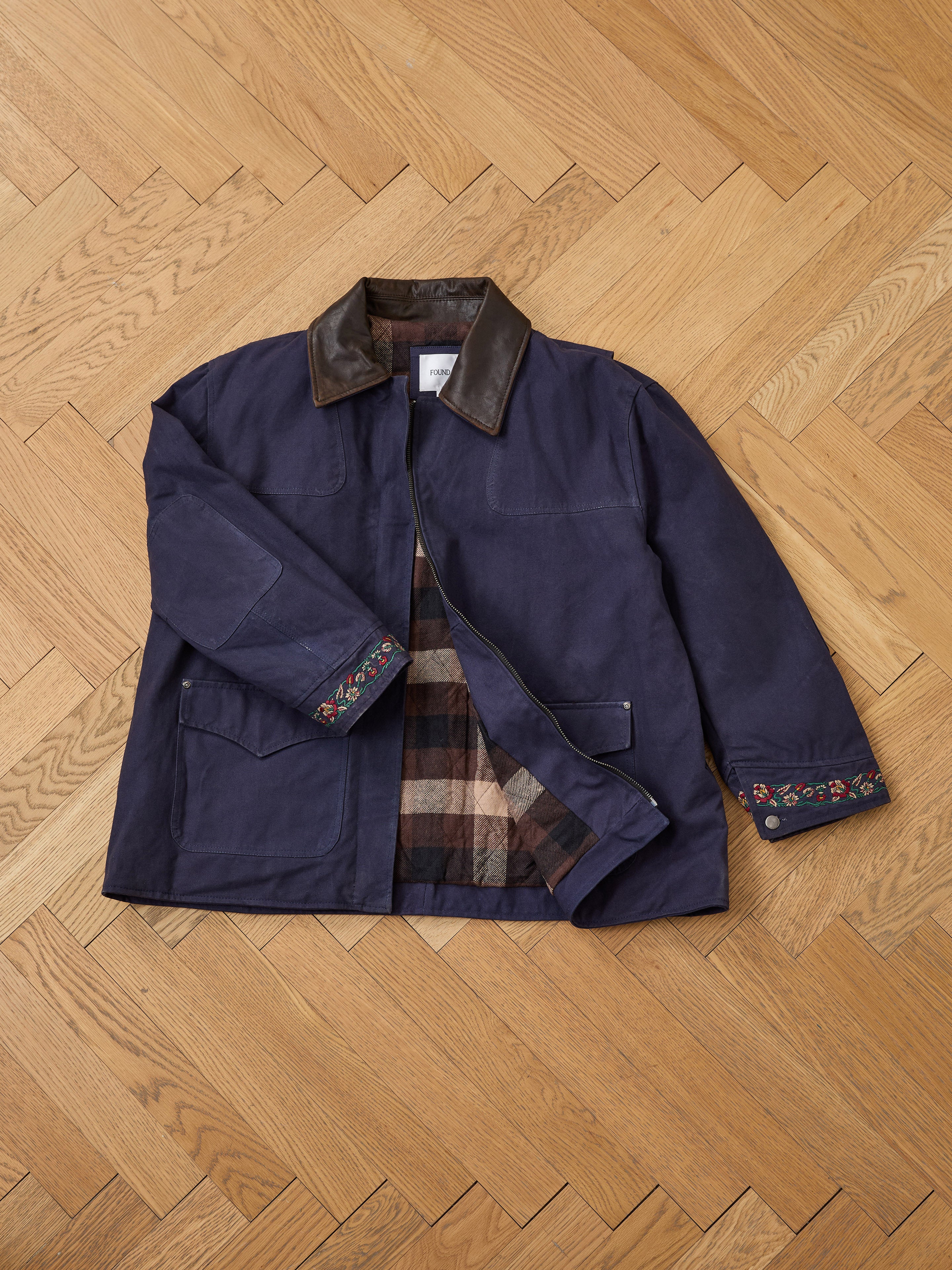 The Found Canvas Distressed Barn Jacket, a unisex relaxed-fit navy piece with a leather collar, plaid lining, front pockets, and floral cuff embroidery, is shown laid flat on a wooden herringbone floor.