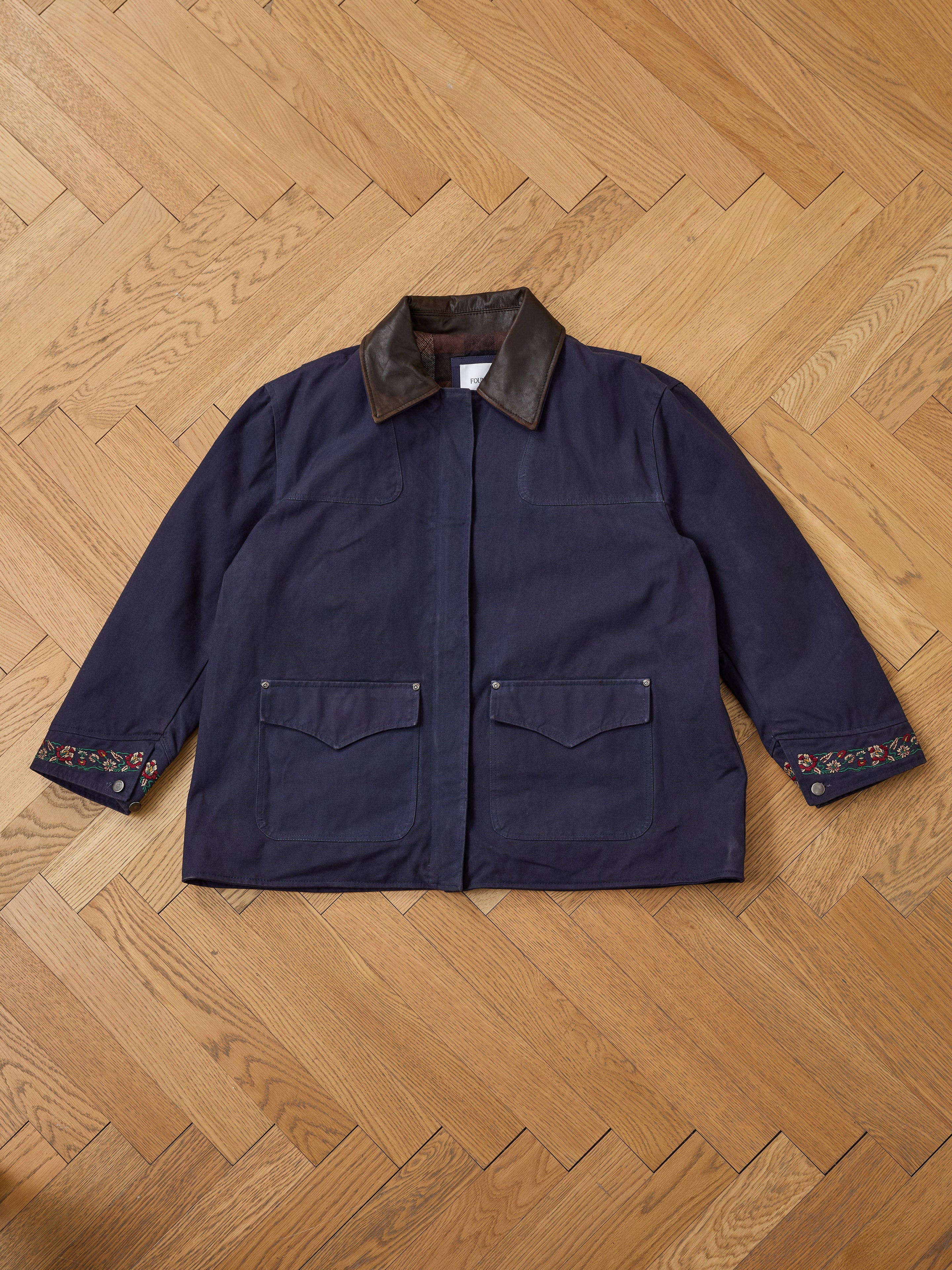 The Found Canvas Distressed Barn Jacket, featuring a relaxed unisex fit and distressed leather collar, is displayed on a wooden floor.