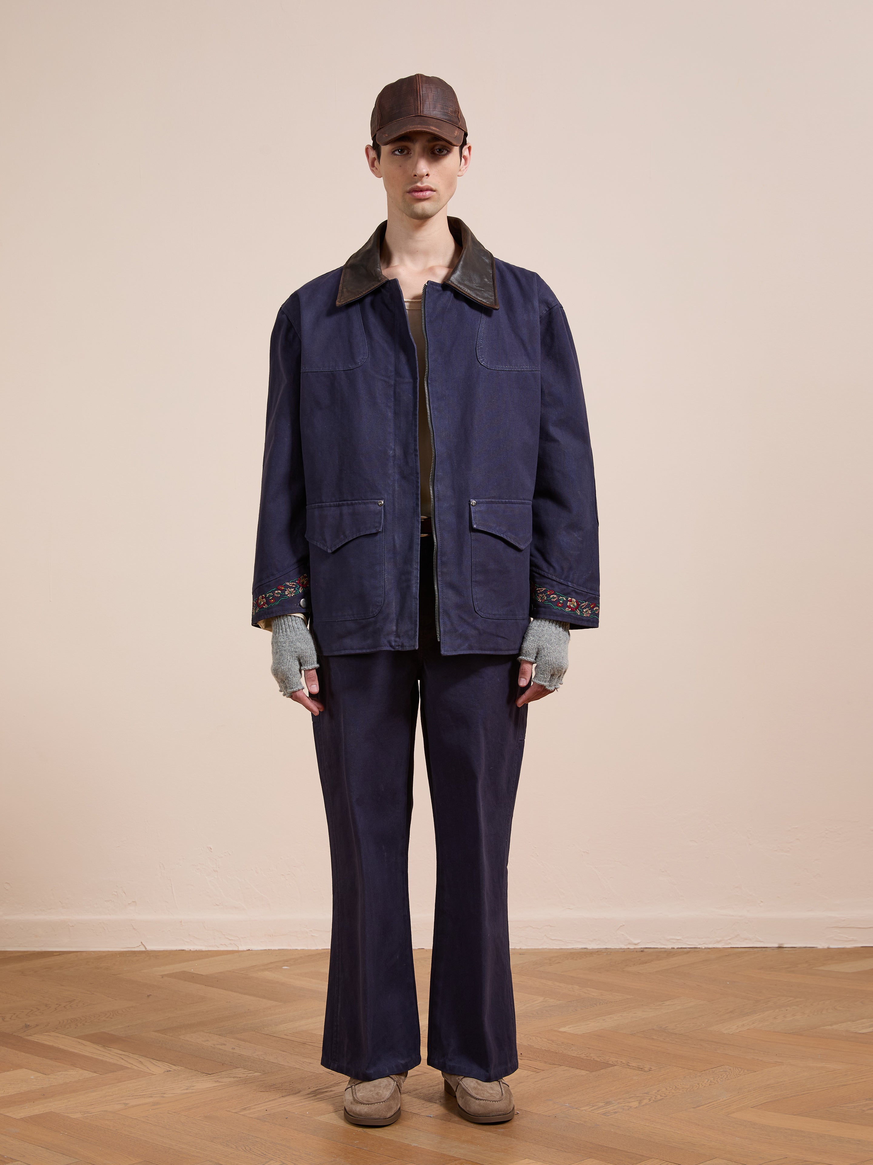 Indoors, a person wears the Found Canvas Distressed Barn Jacket with a worn leather collar, matching pants, brown cap, fingerless gloves, and beige shoes. They stand on a wooden floor against a plain beige wall.