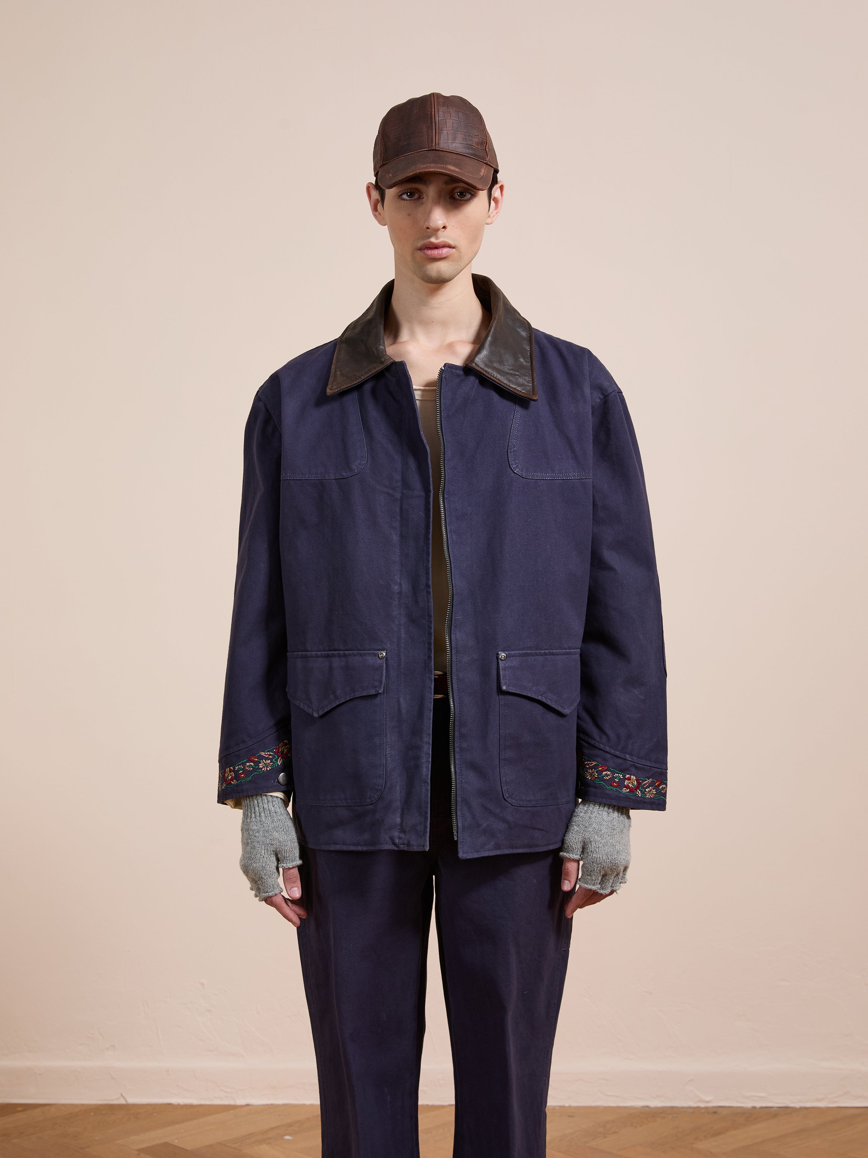 A person stands against a plain background wearing Found's Canvas Distressed Barn Jacket with a distressed leather collar, matching unisex relaxed pants, a brown cap, gray fingerless gloves, and a neutral expression.
